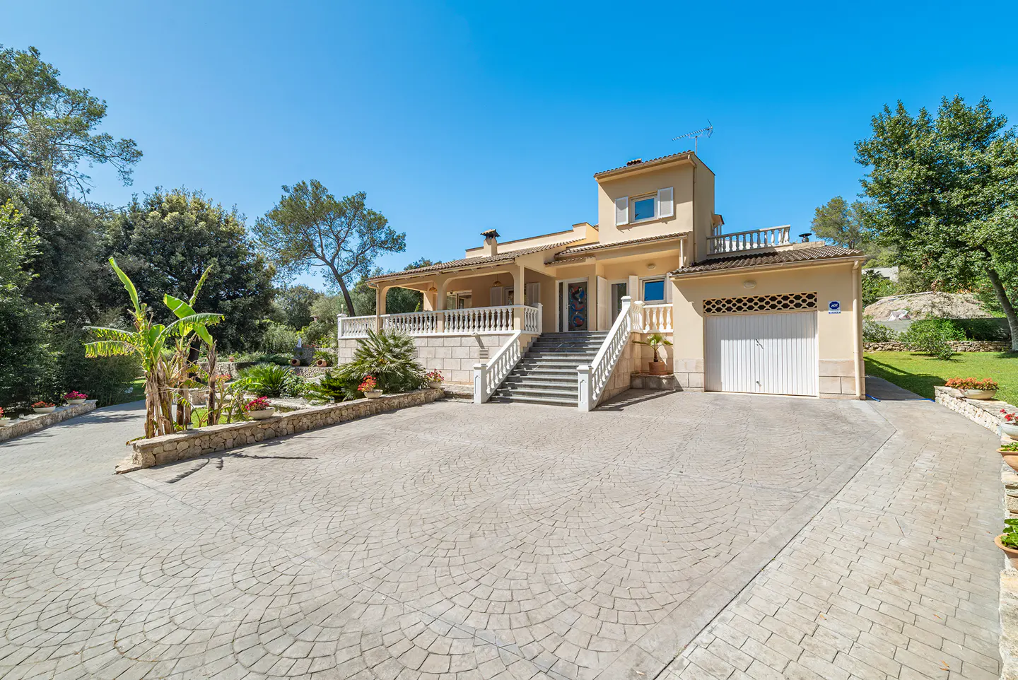 Exterior view of a two-story beige house with a white garage door, stone steps, and a patterned driveway under a clear blue sky.