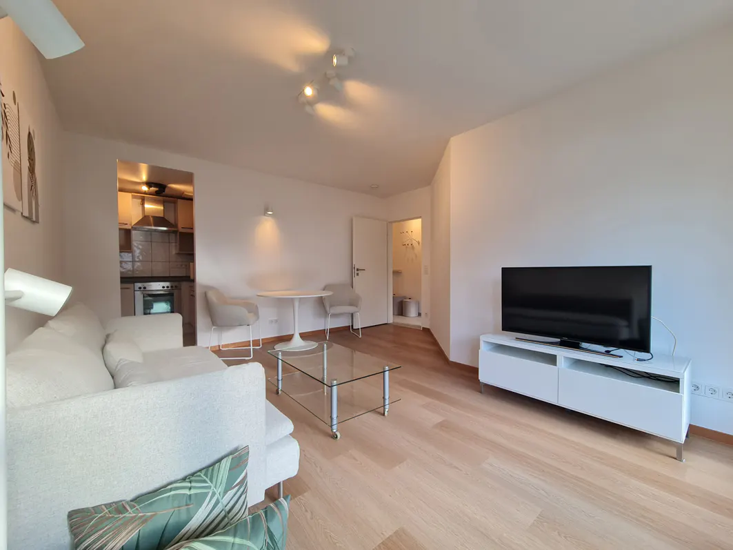 Bright living room with a white sofa, glass coffee table, TV on a white stand, and a view into a modern kitchen.