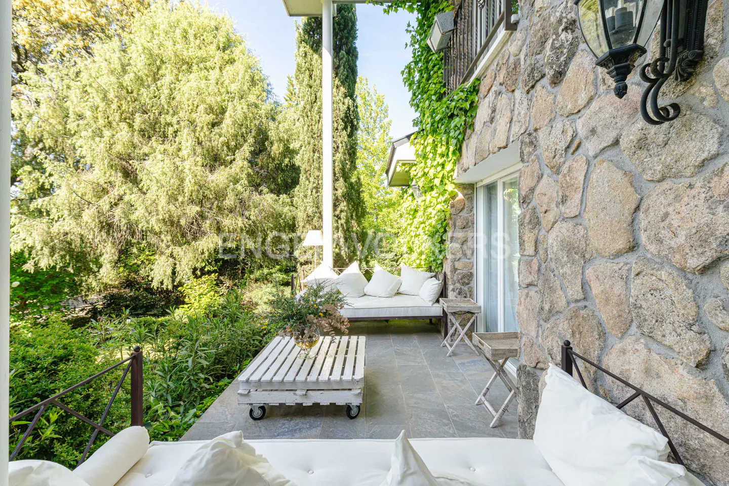 Outdoor patio with stone wall, white cushions, and pallet coffee table. Green trees and vines surround the space.