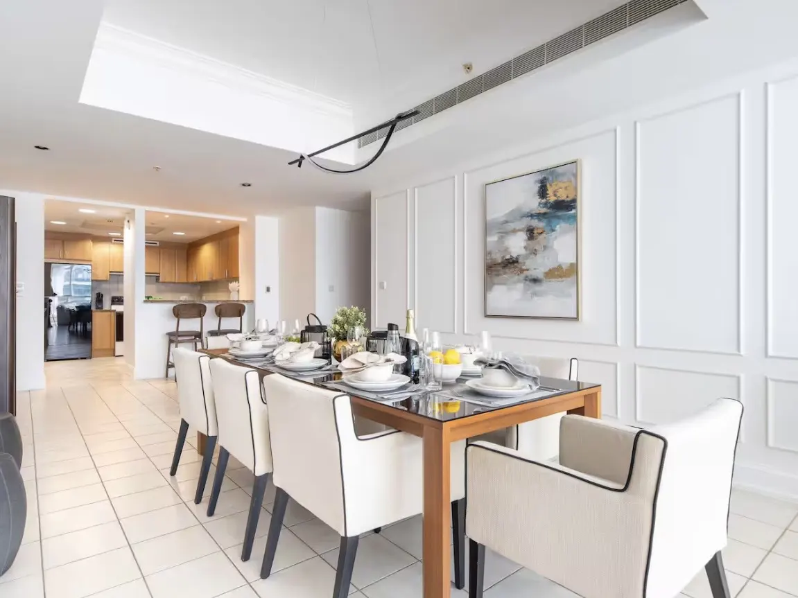 A bright dining room with a long table set for a meal, surrounded by white chairs. An open kitchen is visible in the background.
