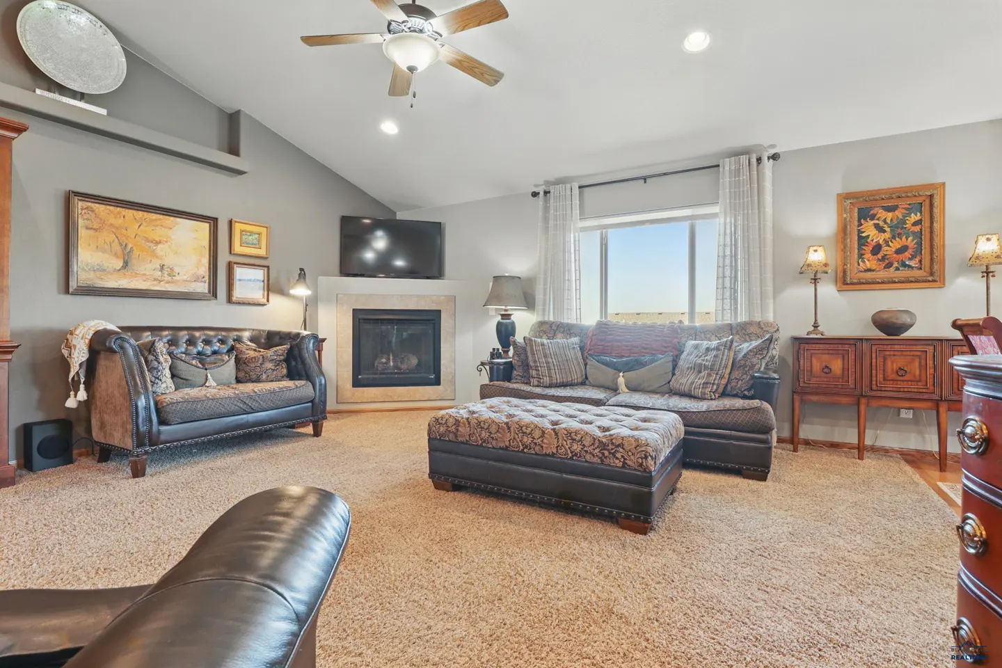 Living room with gray walls, carpet, leather sofa, patterned ottoman, fireplace, and sunflower wall art.