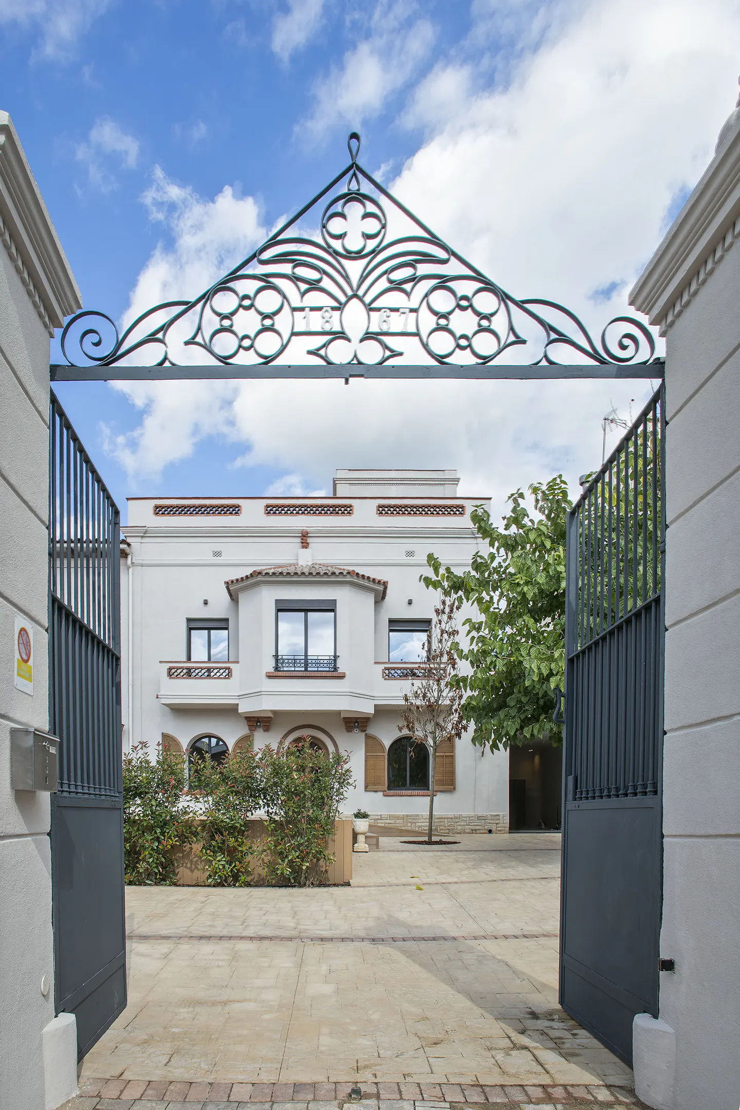 View of a white house through an open black metal gate with decorative top. Blue sky with clouds above.