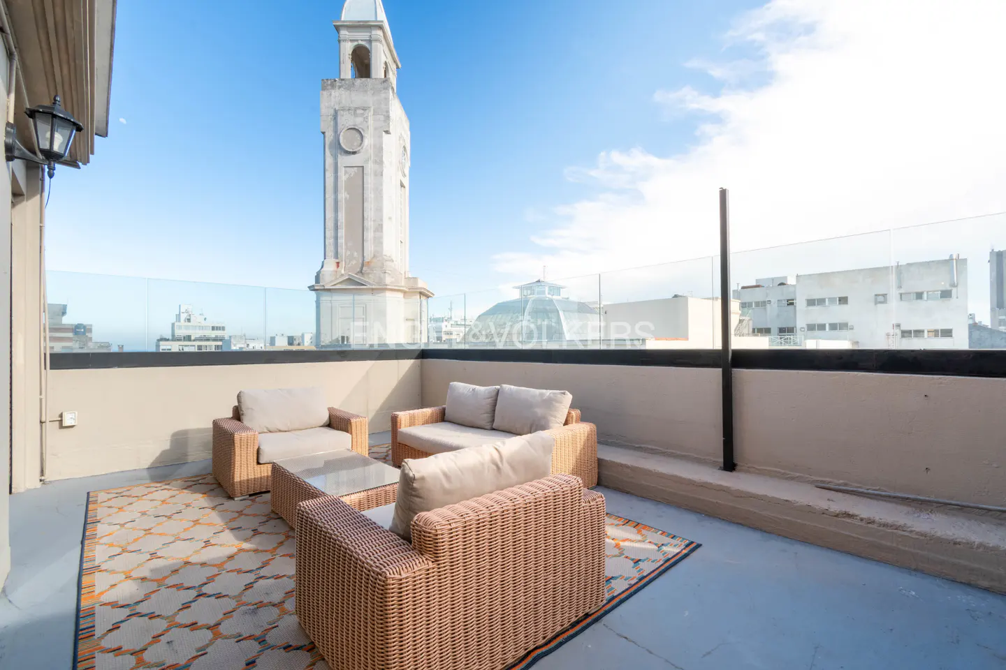 Rooftop patio with wicker furniture, glass railing, and city views. A tall clock tower rises in the background against a blue sky.