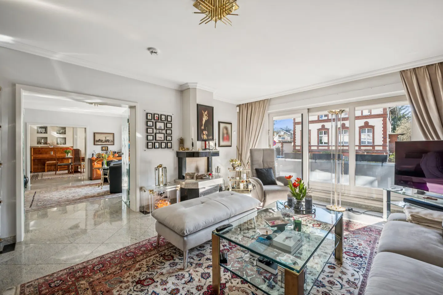 Bright living room with a fireplace, gray sofa, glass coffee table, and a view of a building through a large window.