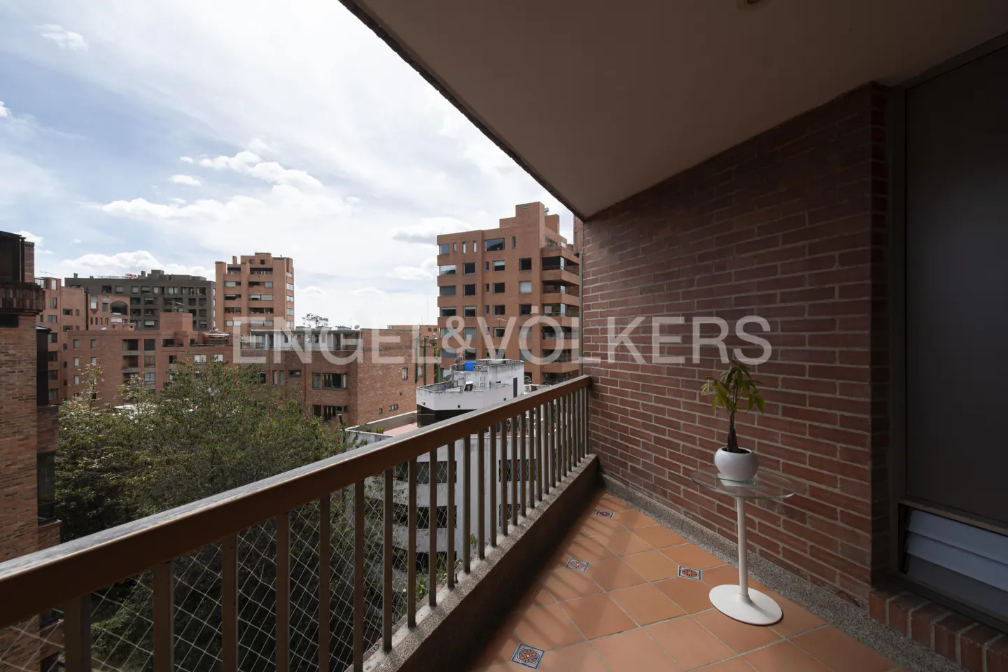 Balcony view with brick walls and brown tile floor. A plant sits on a white stand. Buildings and sky are visible beyond the railing.