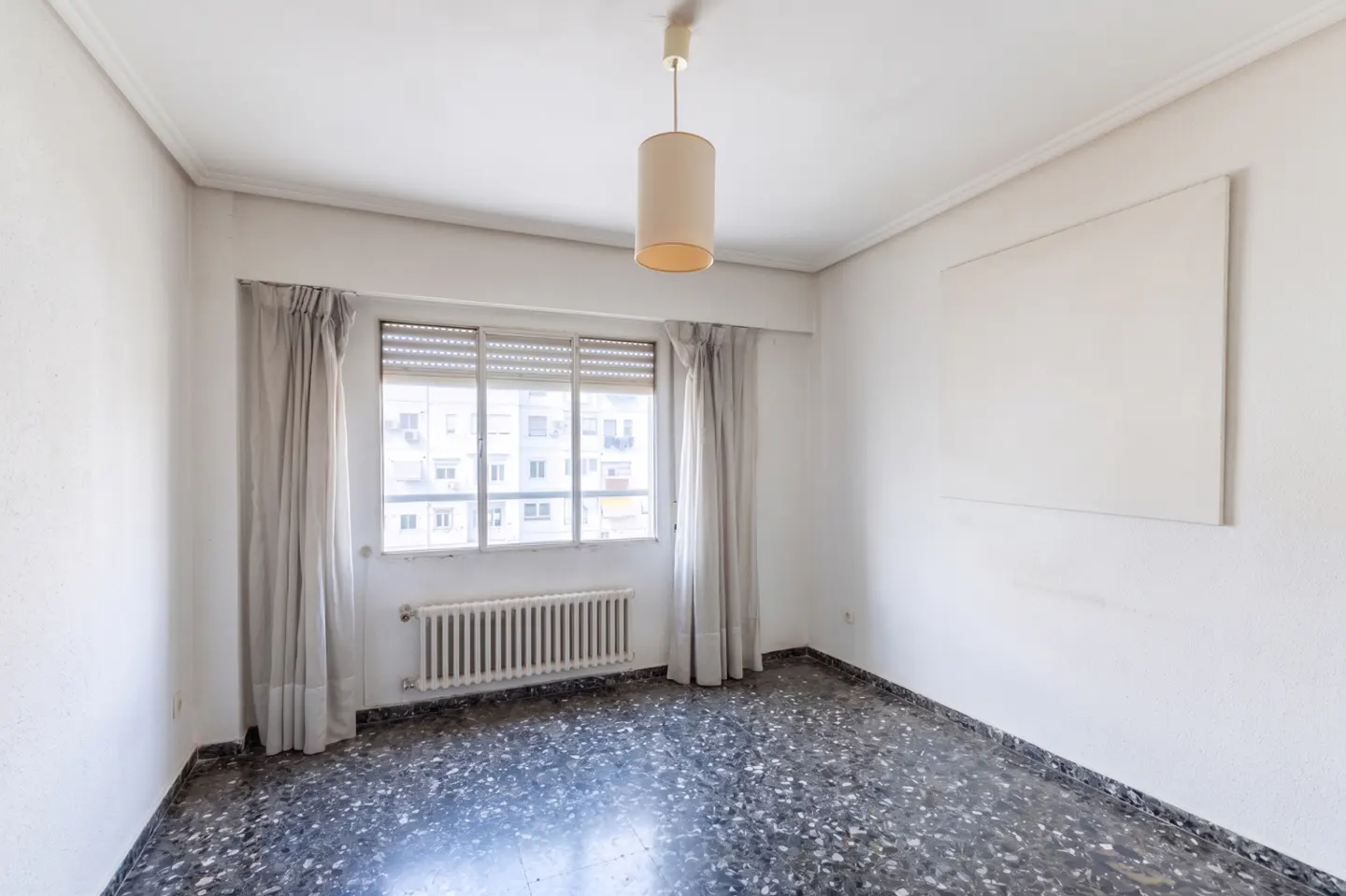 Empty room with white walls, a window with blinds and curtains, a radiator, and a dark speckled floor. A cylindrical lamp hangs from the ceiling.
