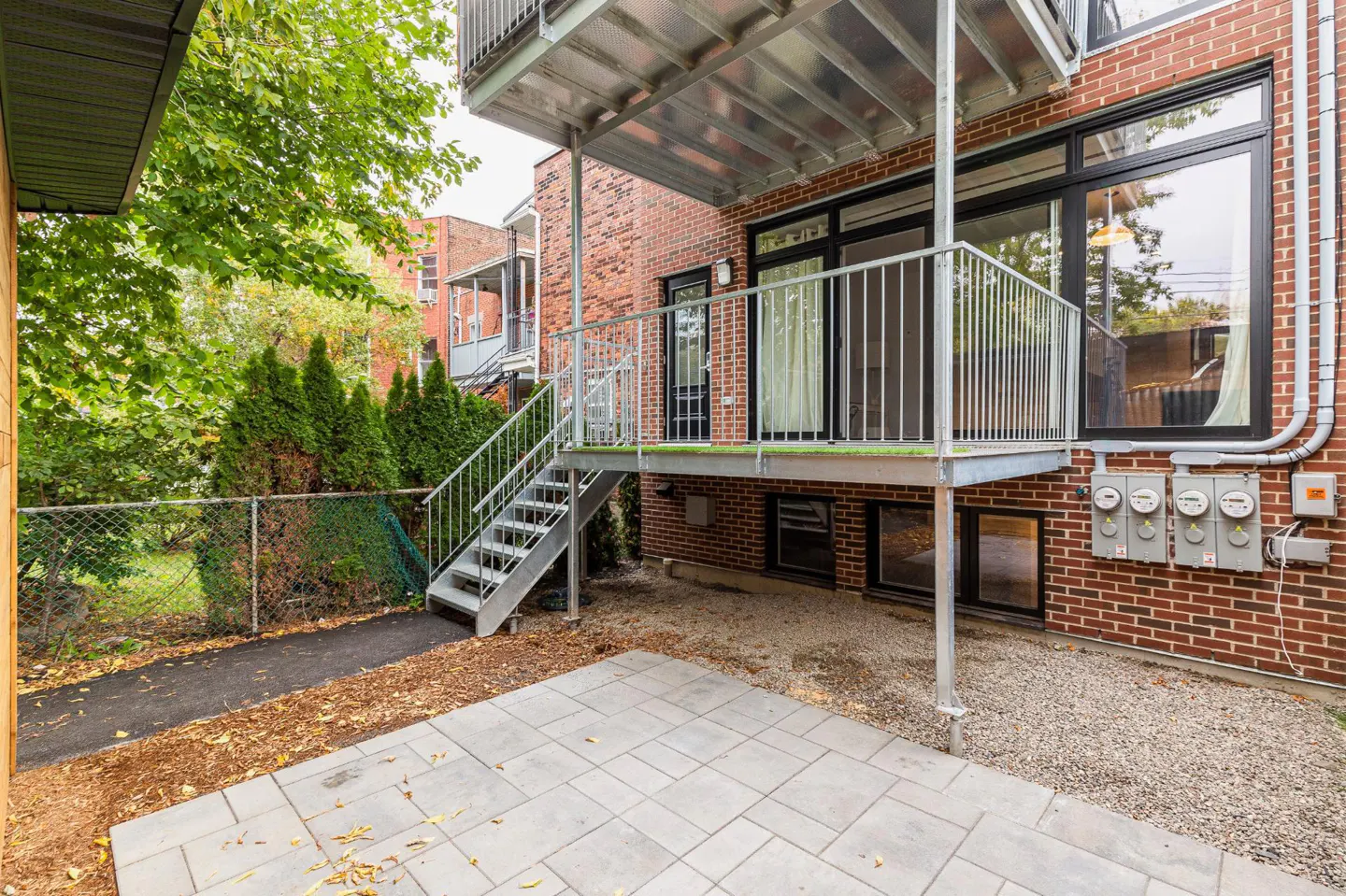 Brick building exterior with a gray stone patio, metal stairs to a balcony, and green trees.