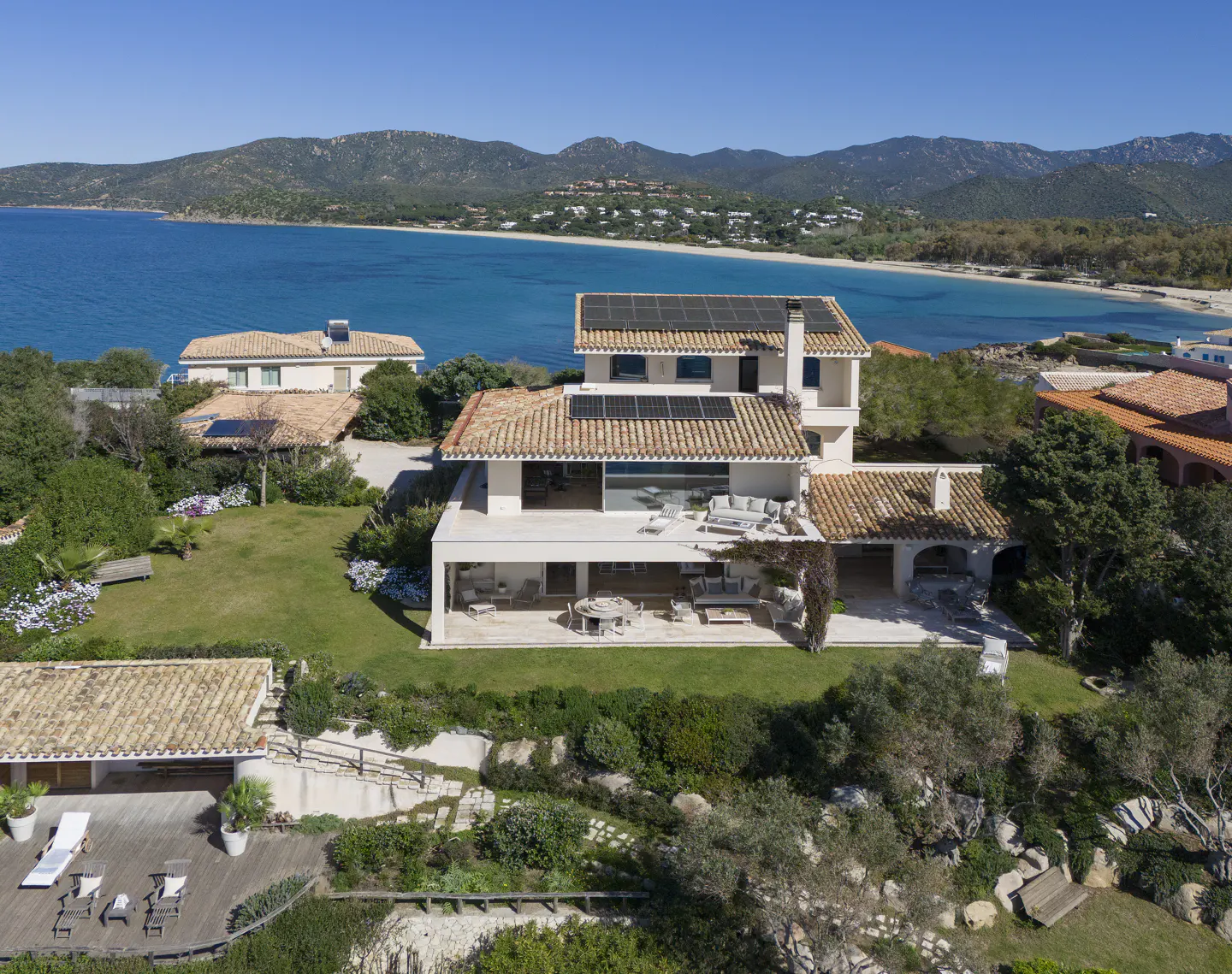 Aerial view of a modern white house with a terracotta roof and solar panels, overlooking a turquoise bay and mountains.