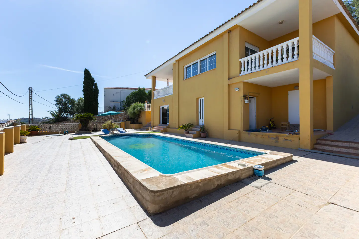 Two-story yellow house with a blue swimming pool and tiled patio on a sunny day.