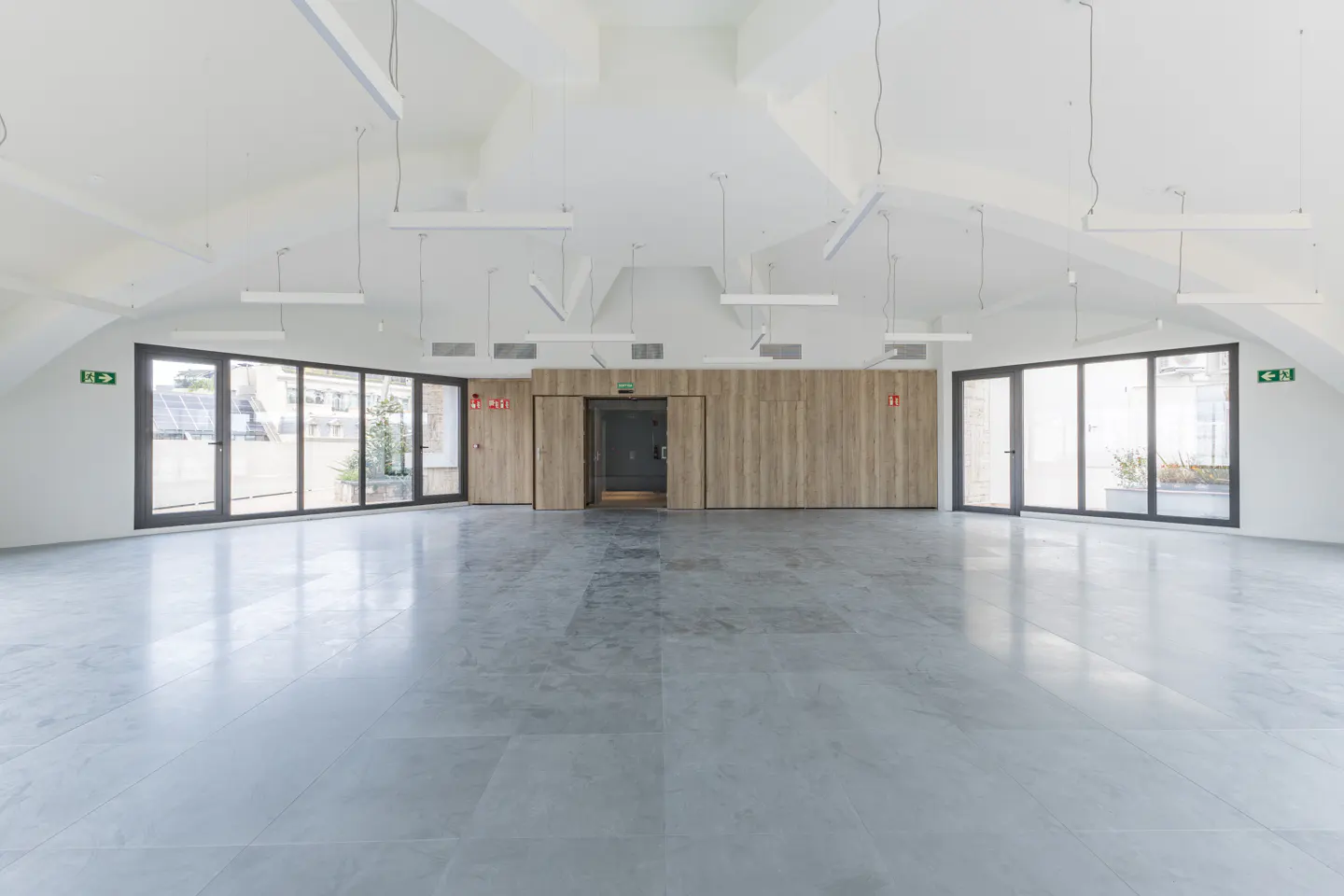 Bright, empty room with gray tile floor, white walls, and a vaulted ceiling. Large windows and a wood-paneled wall with a door are visible.