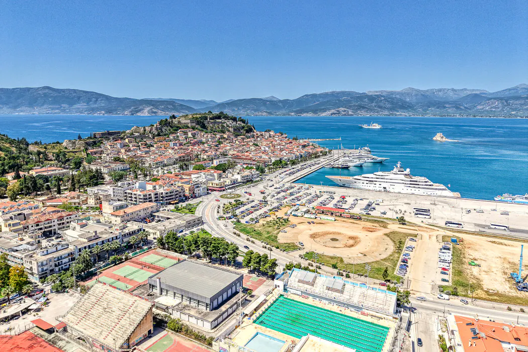 Aerial view of Nafplio, Greece, featuring red-roofed buildings, a harbor with yachts, and mountains in the background under a clear blue sky.