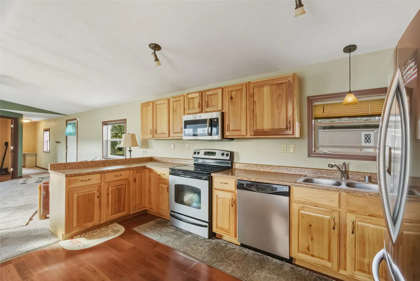 A kitchen with light wood cabinets, stainless steel appliances, and a window with blinds.