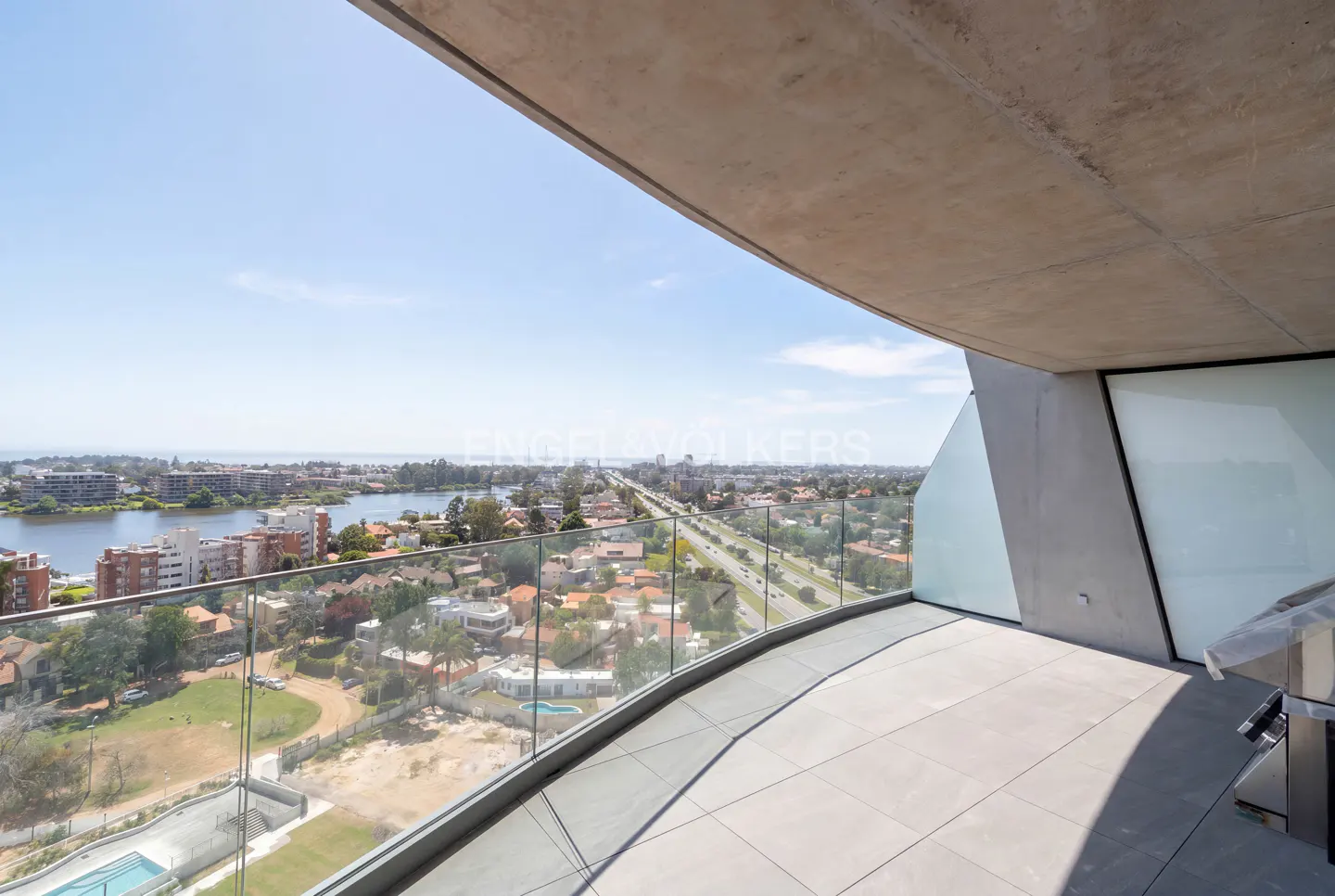 Modern balcony with concrete ceiling and glass railing overlooking a suburban landscape, river, and distant city skyline.