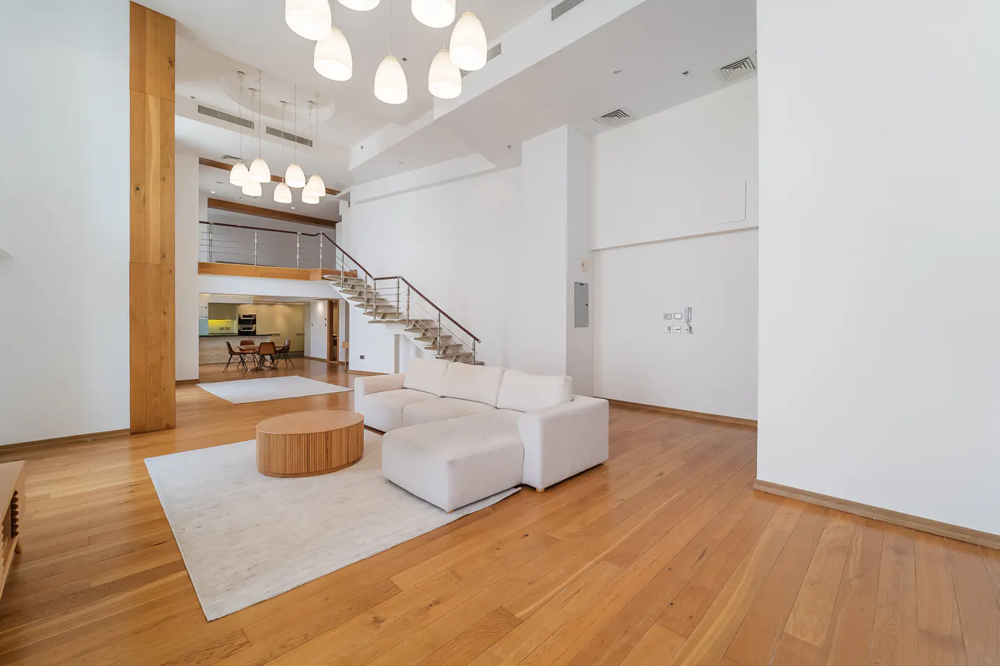 Bright, open-plan living room with hardwood floors, white walls, and a staircase leading to a mezzanine level. A white sofa and round wooden table sit on a cream rug.