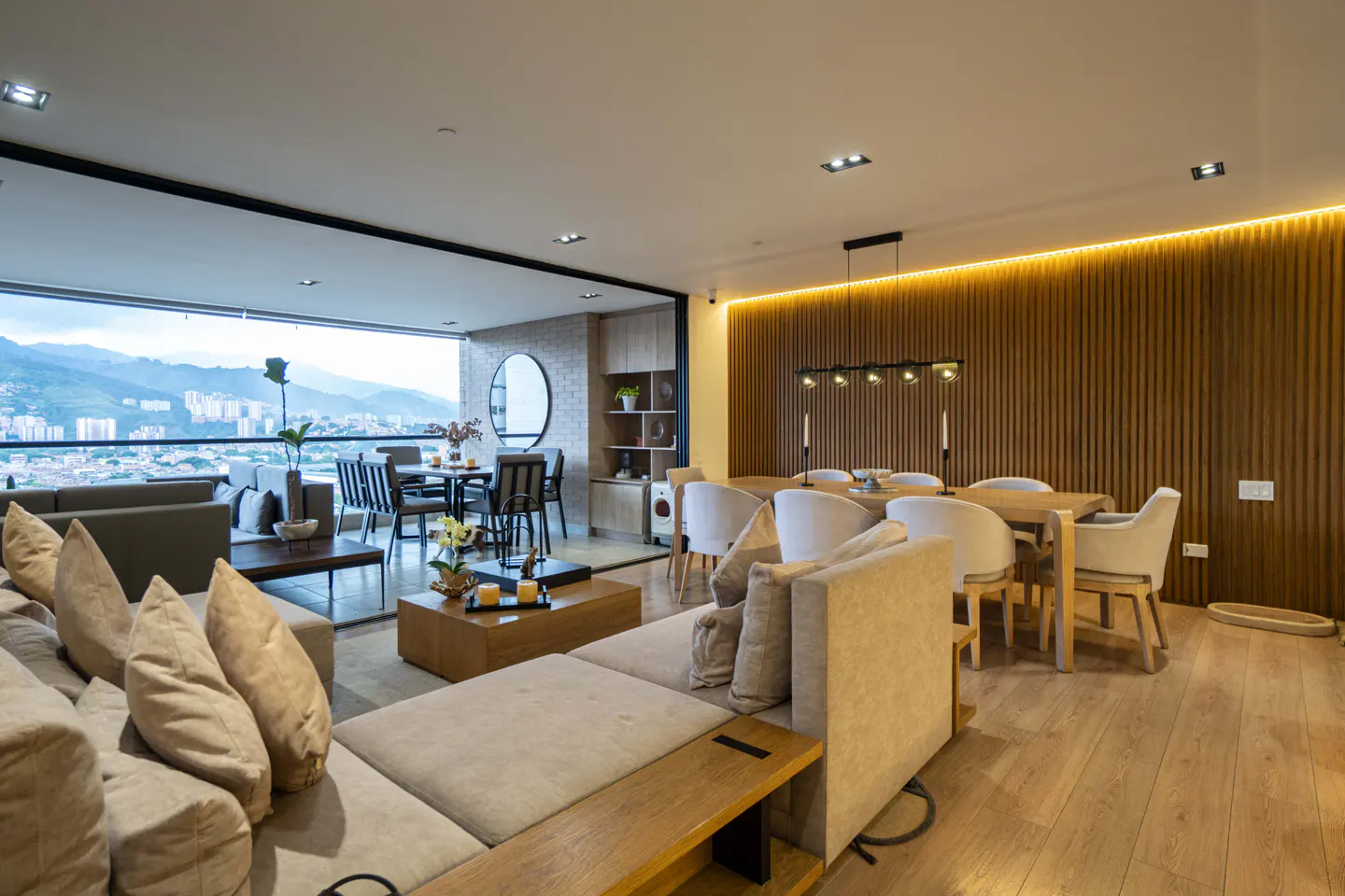 Bright, modern living and dining area with a beige sectional sofa, wood floors, and a city view from a large window.