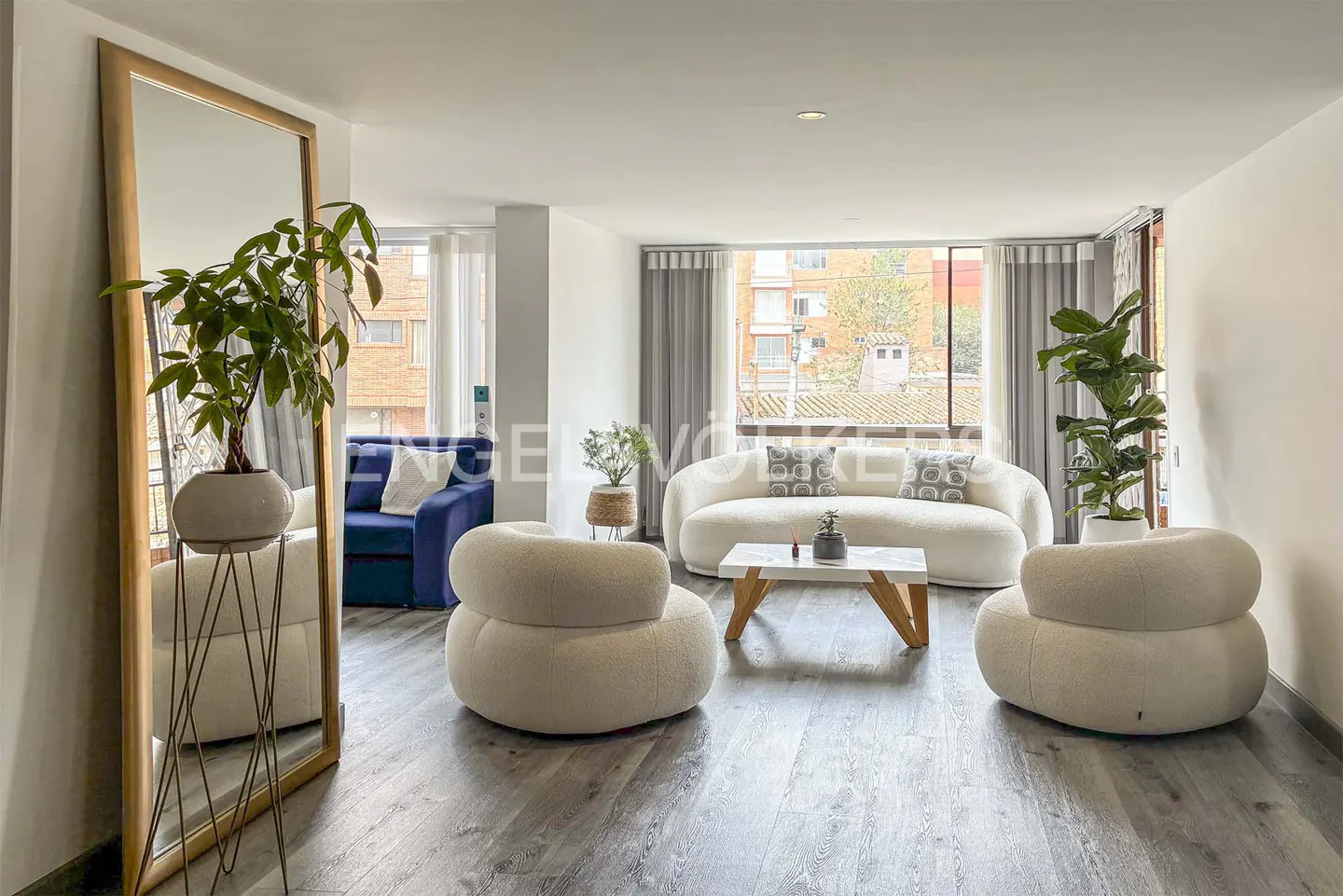 Bright living room with gray wood floors, white walls, and large windows. Features a white sofa, two round chairs, and a blue loveseat reflected in a mirror.