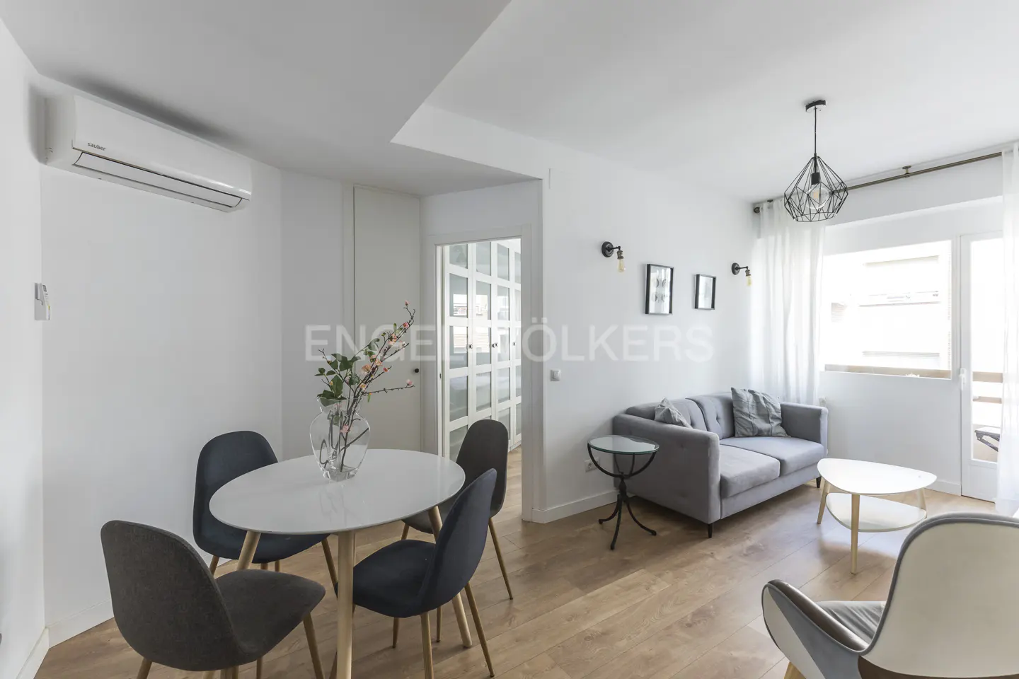 Bright living room with wood floors, white walls, and modern furniture. A round table with chairs sits near a gray sofa and balcony.