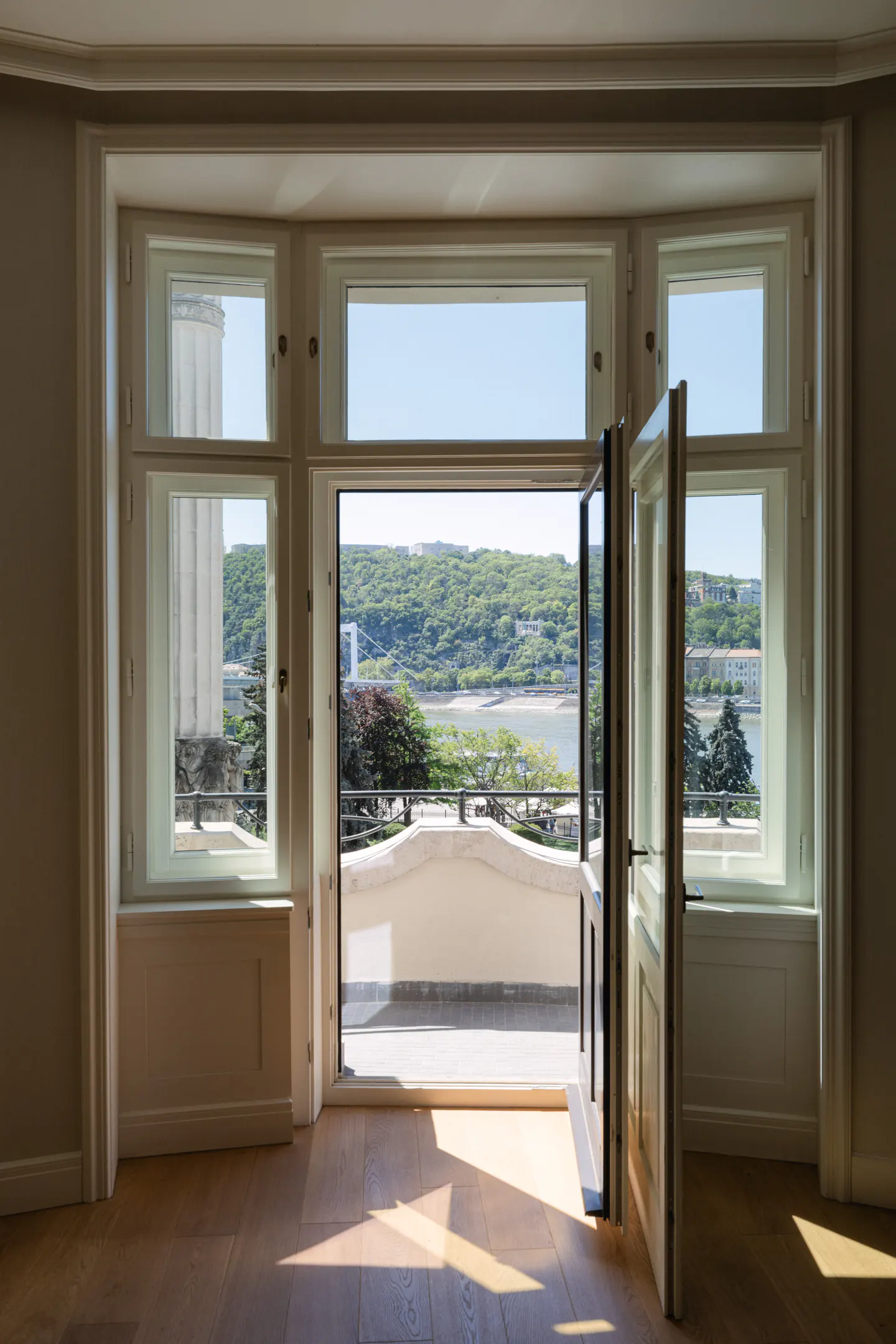 Bright room with open French doors leading to a balcony overlooking a river and green hills. Hardwood floors and white trim.
