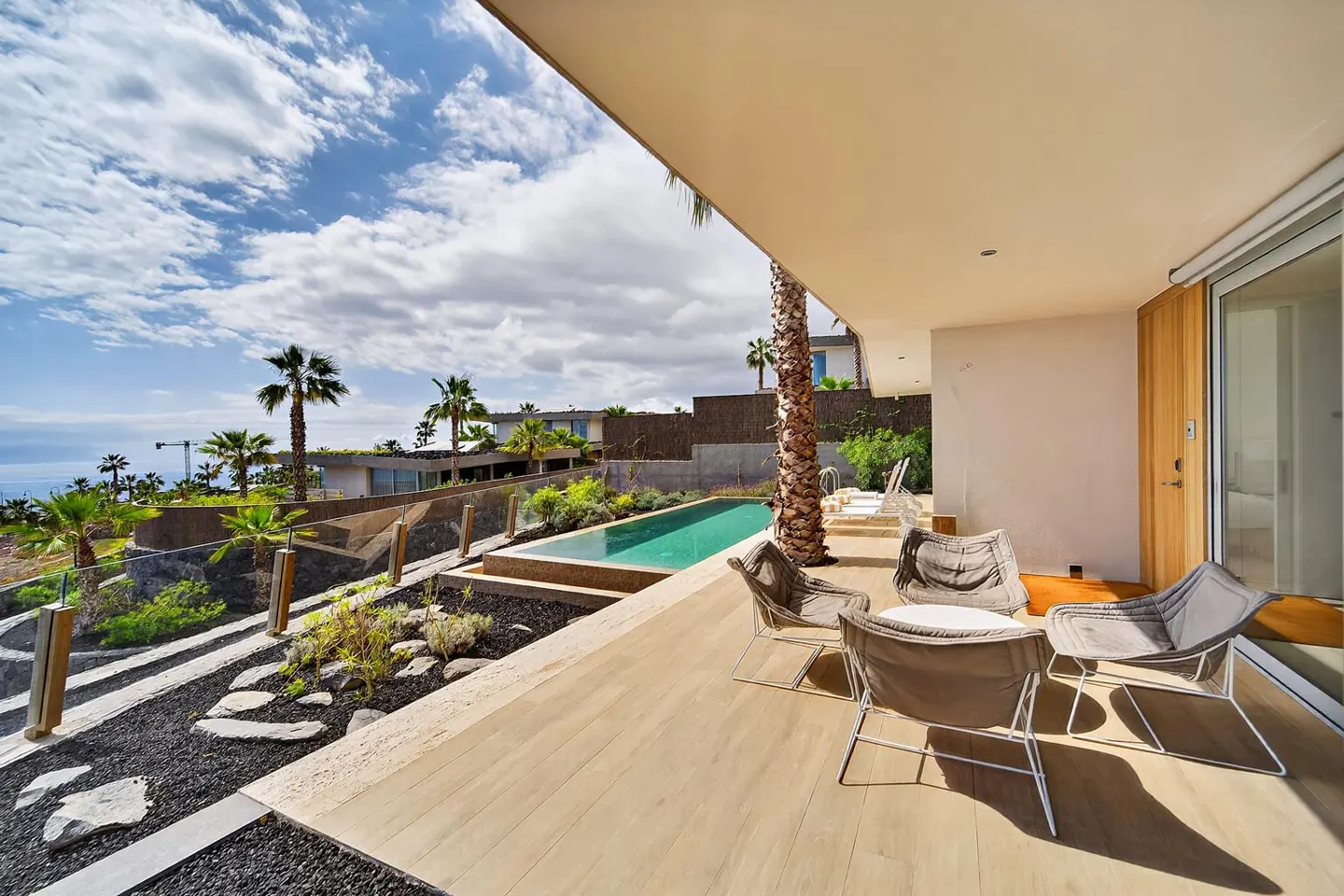Outdoor patio with four chairs, a small table, and a pool overlooking the ocean and palm trees under a cloudy sky.