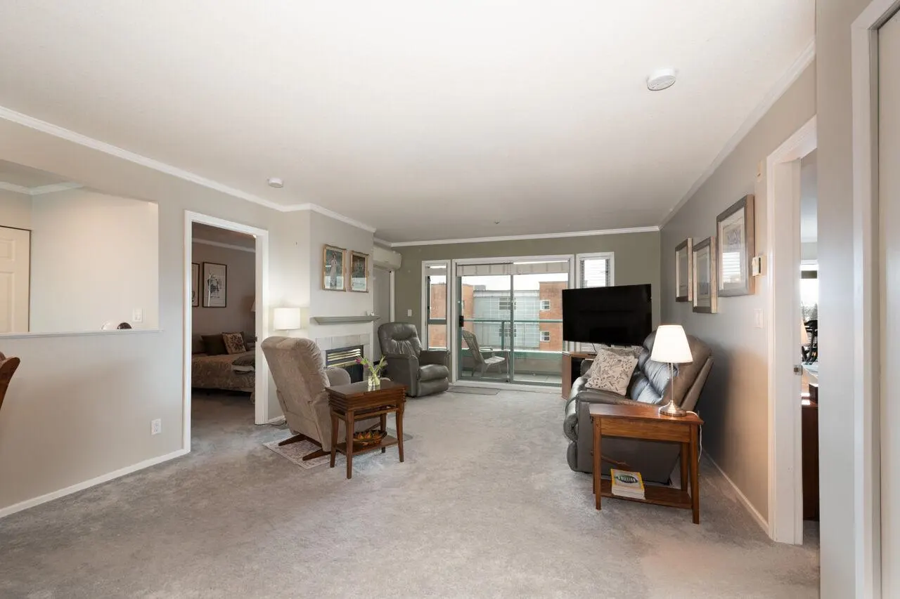 Living room with gray carpet, walls, and furniture. A fireplace, TV, and balcony are also visible.