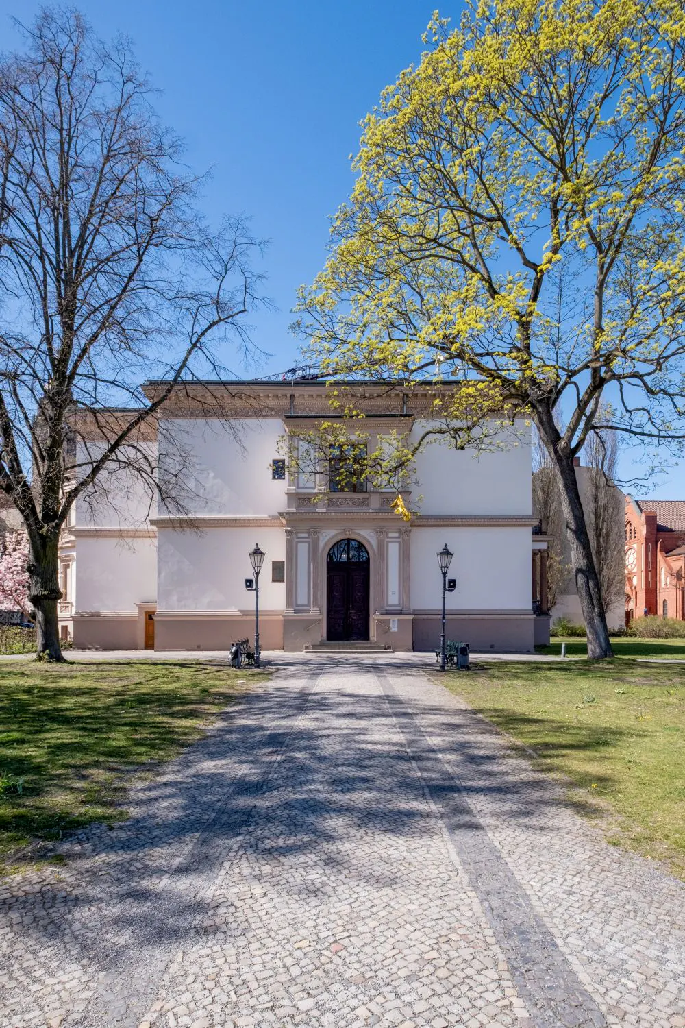 Exterior view of a cream-colored building with a dark wood door, framed by trees and a cobblestone path on a sunny day.