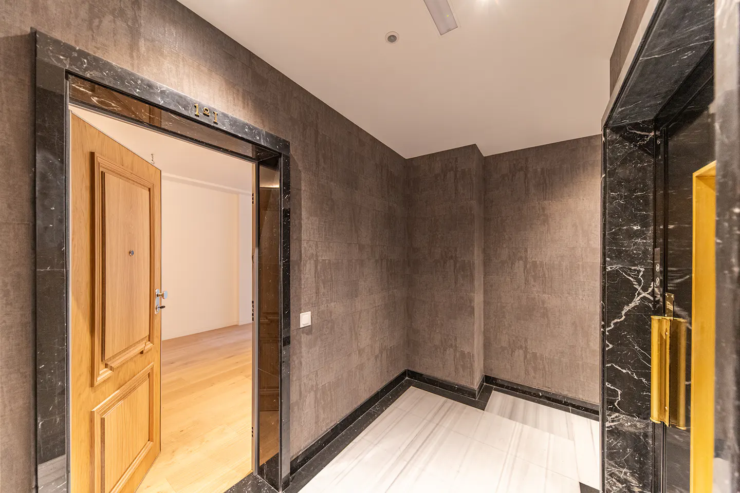 A hallway with dark textured walls, a marble floor, a wooden apartment door labeled 121, and a black marble elevator frame.