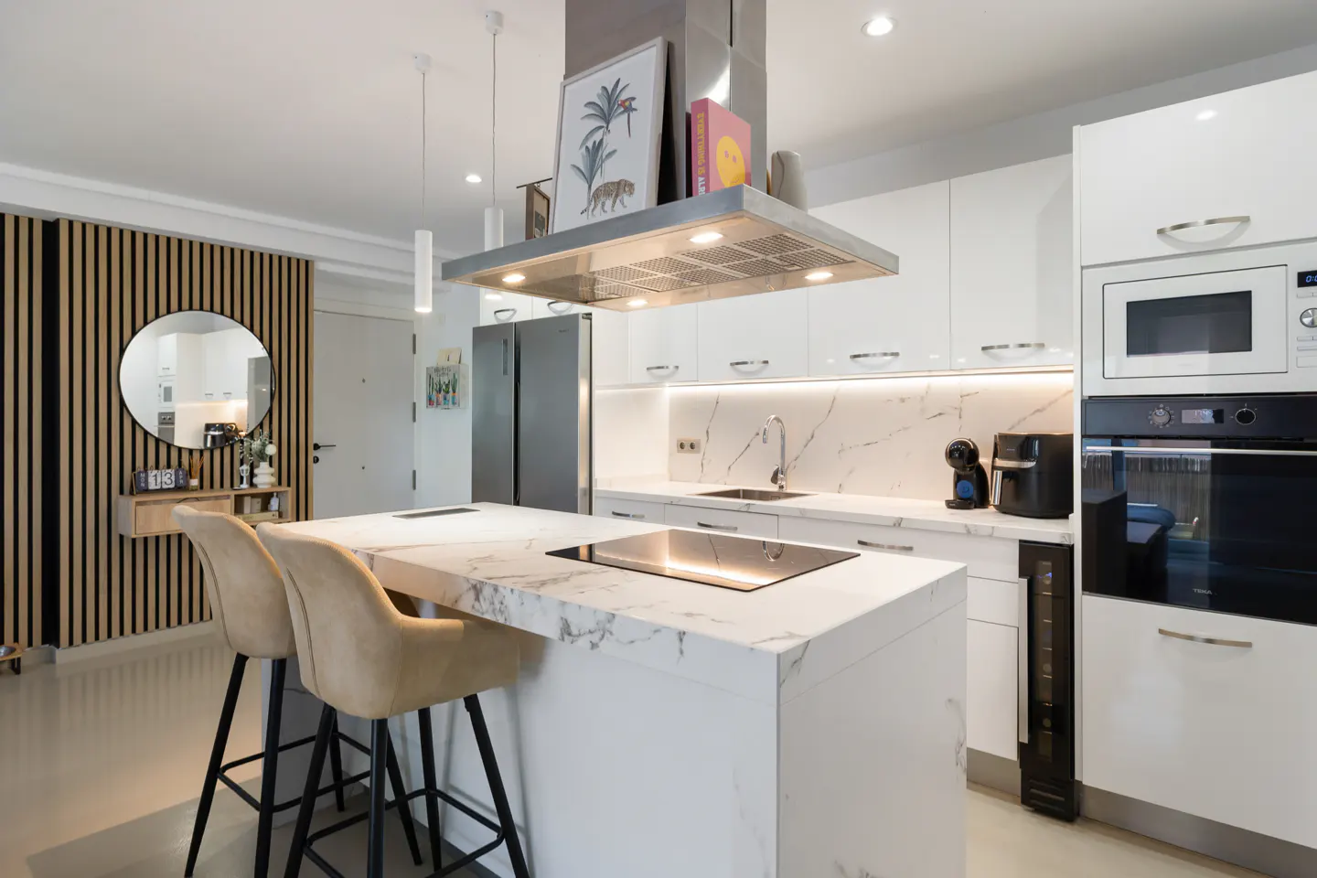 Bright kitchen with white cabinets, marble countertops, and stainless steel appliances. Two beige bar stools face the island.