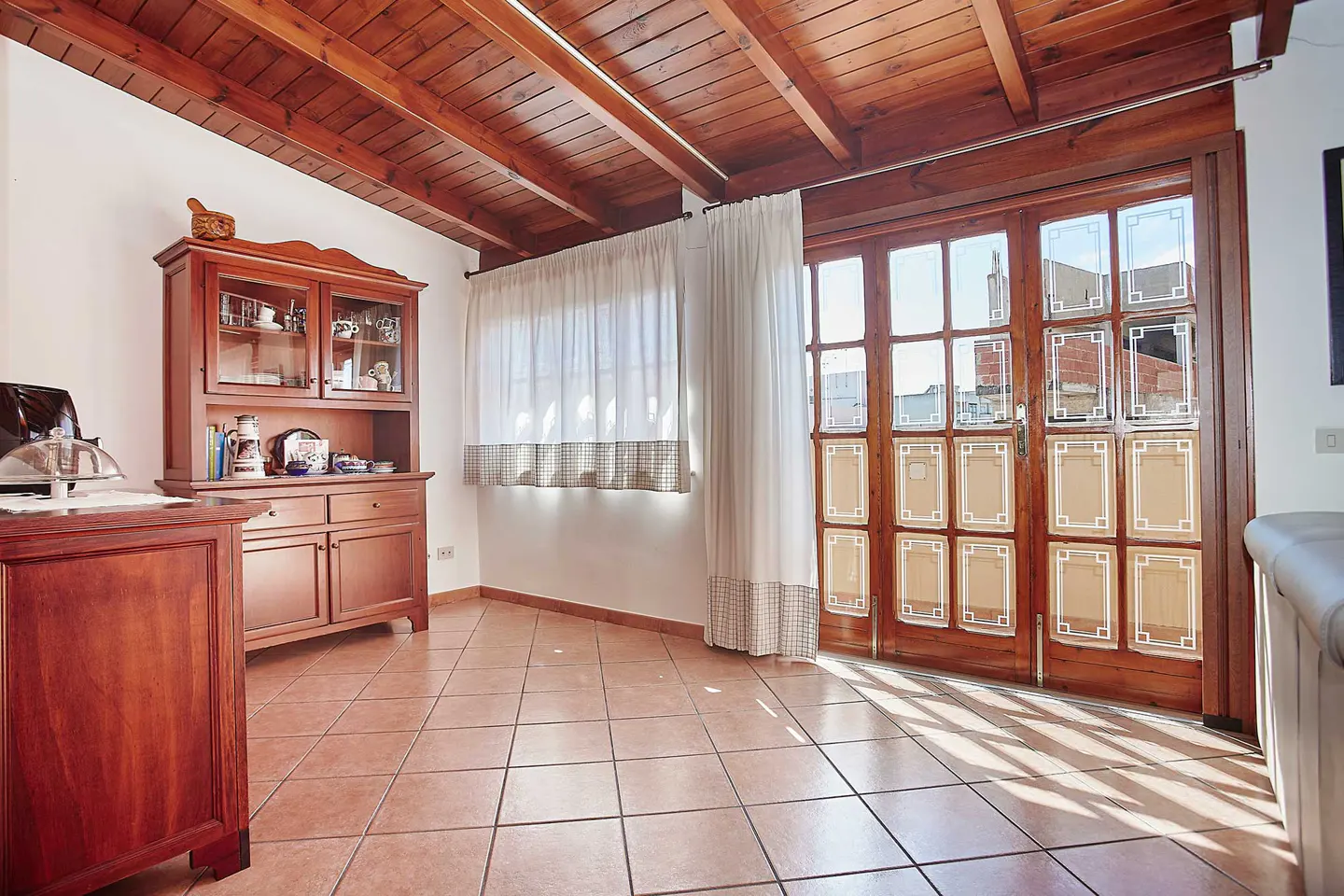 A room with a wood ceiling, tile floor, and a wood hutch. Sunlight streams through a wood-framed glass door with white curtains.