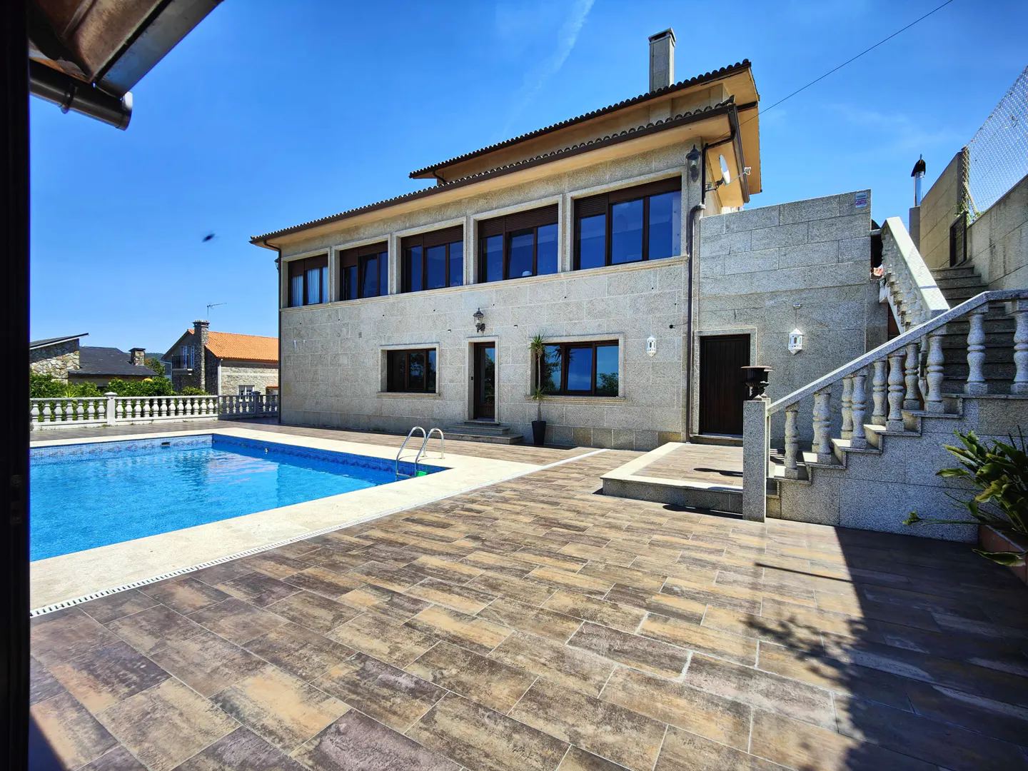 Two-story stone house with a swimming pool, tiled patio, and stone staircase under a clear blue sky.