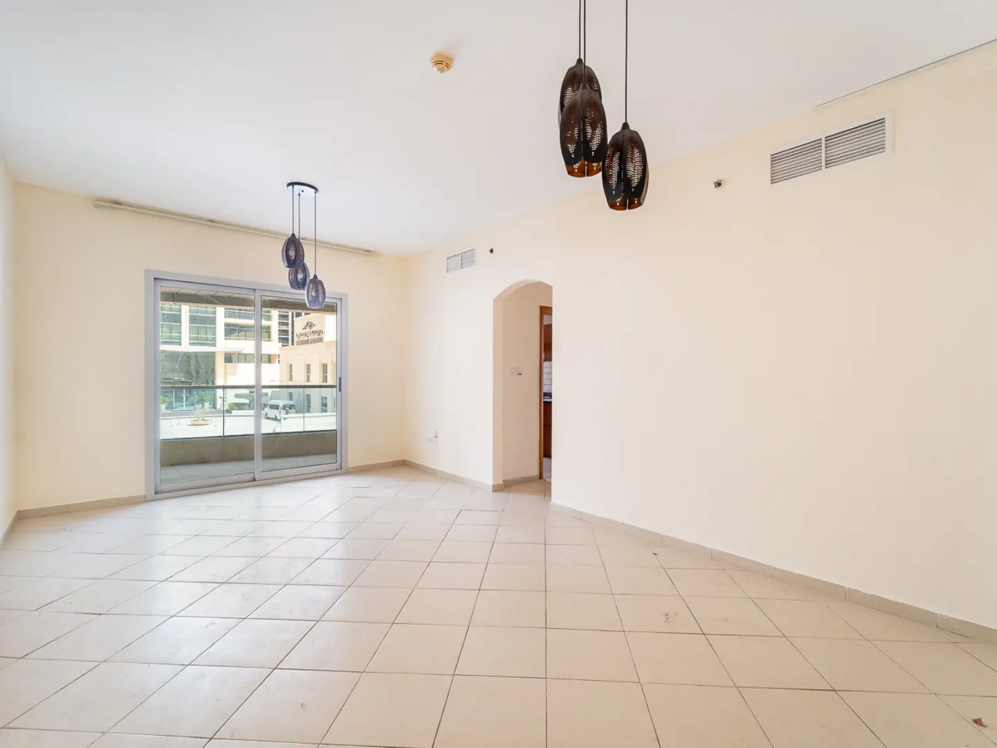 Empty living room with beige walls, tiled floor, sliding glass doors to a balcony, and two sets of pendant lights.