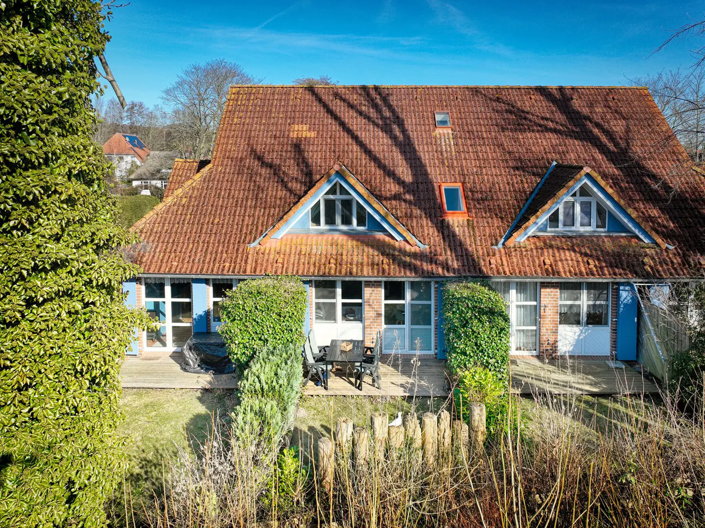 Exterior of a two-story house with a red tile roof, blue trim, and a wooden deck with a table and chairs. Green foliage surrounds the house.