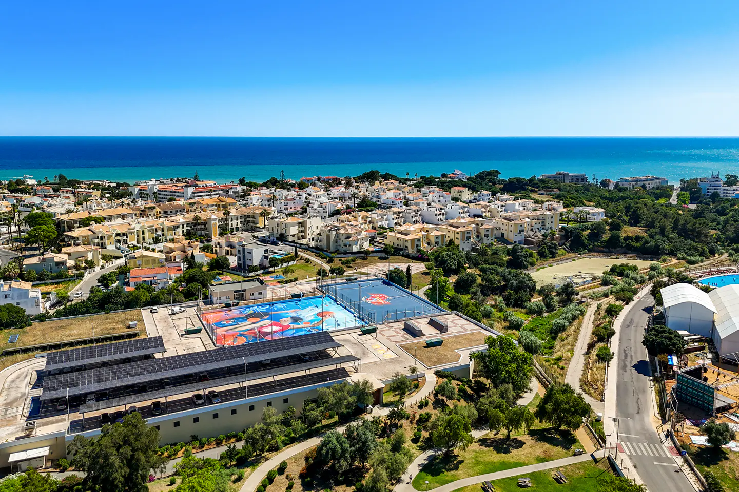 Aerial view of a coastal town with a colorful basketball court, solar-paneled parking, and the blue ocean in the background.