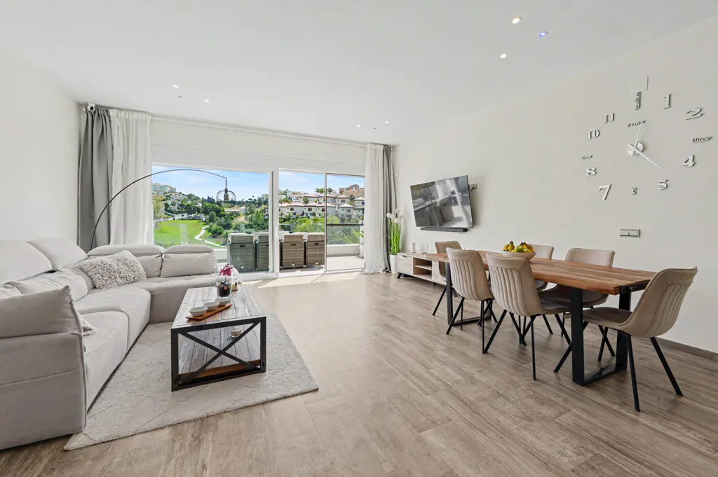 Bright living room with a gray sectional sofa, wood coffee table, dining table with six chairs, and a large clock on the wall. Balcony with a view.