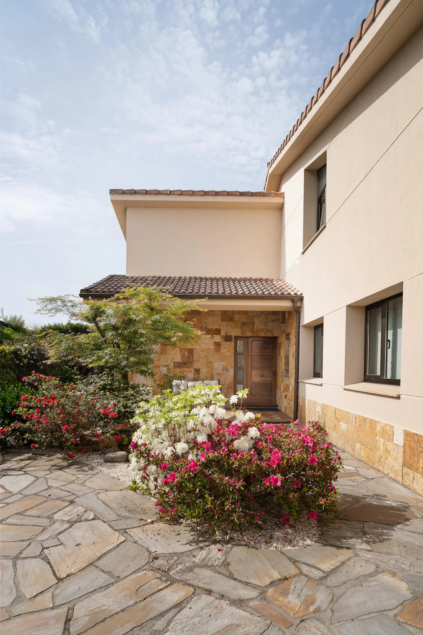Exterior of a beige two-story house with a stone facade, brown roof, and a stone walkway with pink and white flowers.