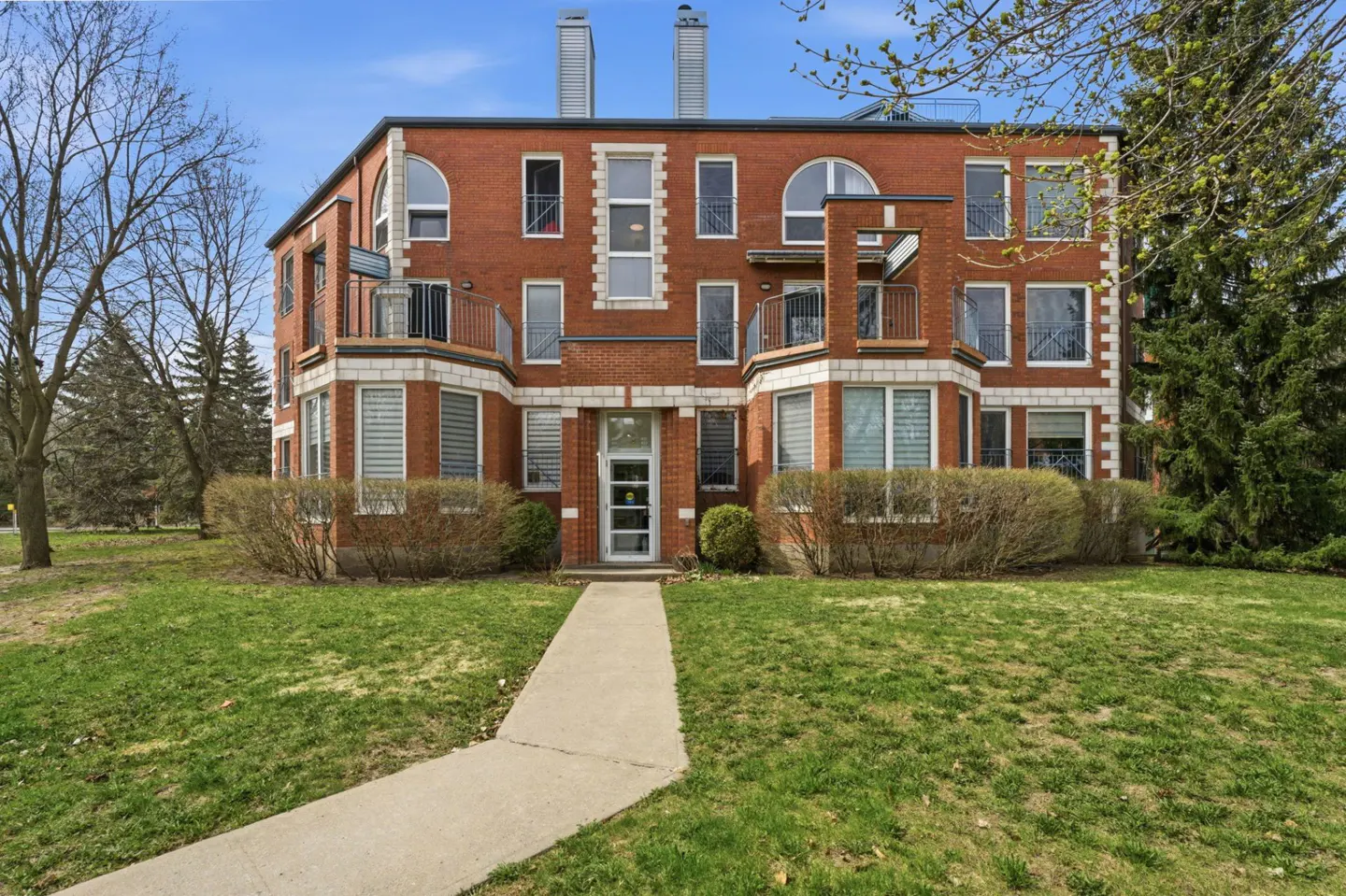 Three-story red brick apartment building with white trim and balconies, set on a green lawn with a concrete walkway.