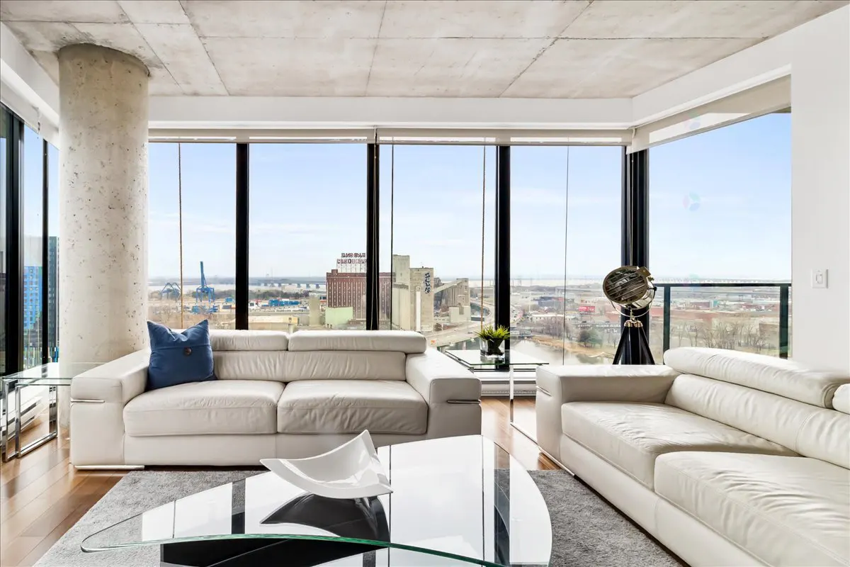 Bright living room with white sofas, glass tables, and floor-to-ceiling windows overlooking a cityscape.