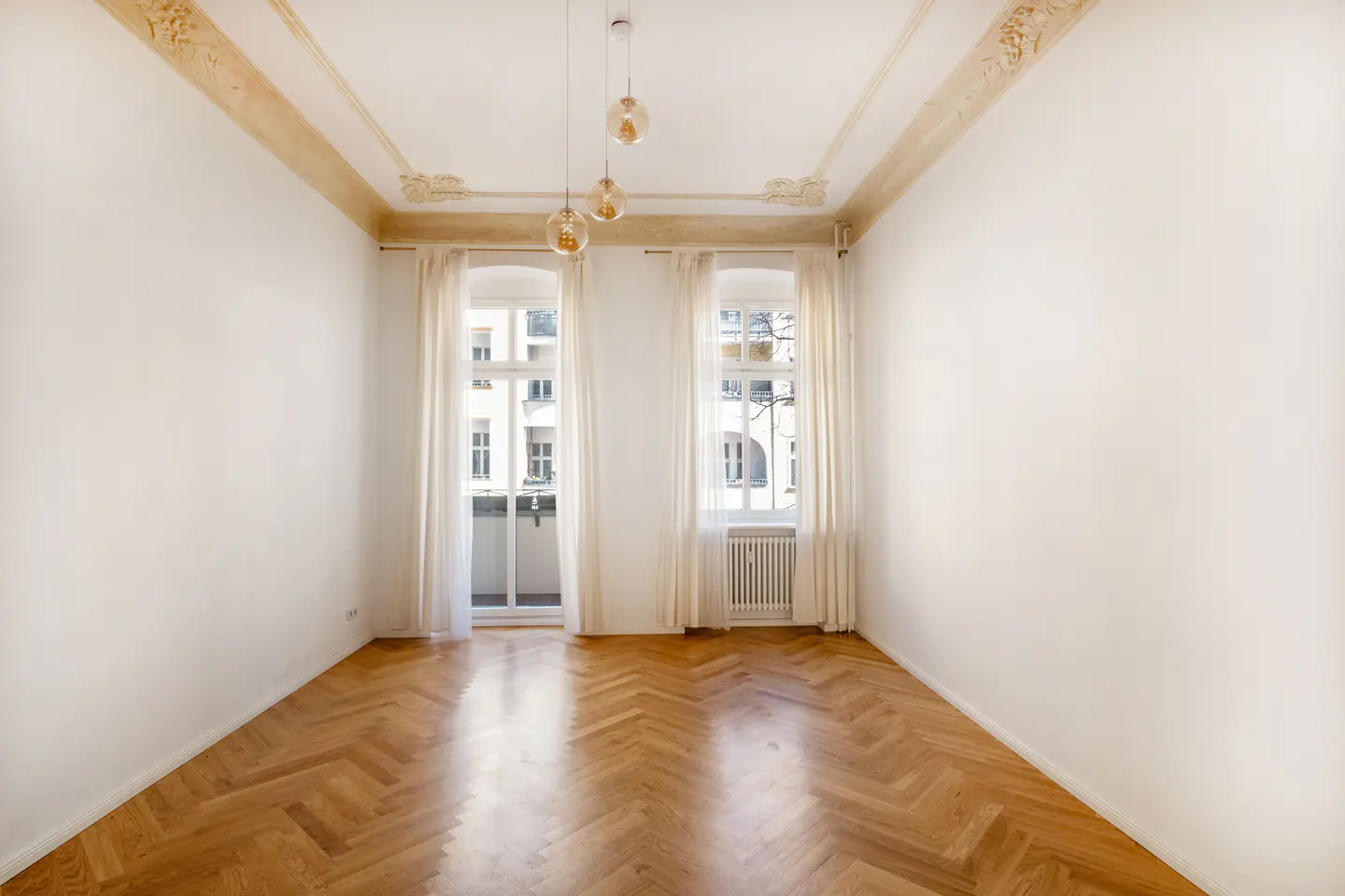 Empty room with herringbone wood floors, white walls, and two windows with white curtains. Three pendant lights hang from the ceiling.