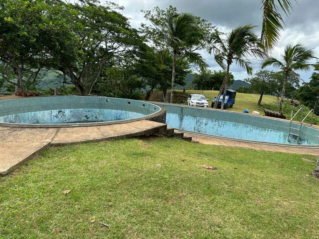 Two empty, light blue pools sit on a grassy hill, with trees, a white car, and a blue vehicle in the background.