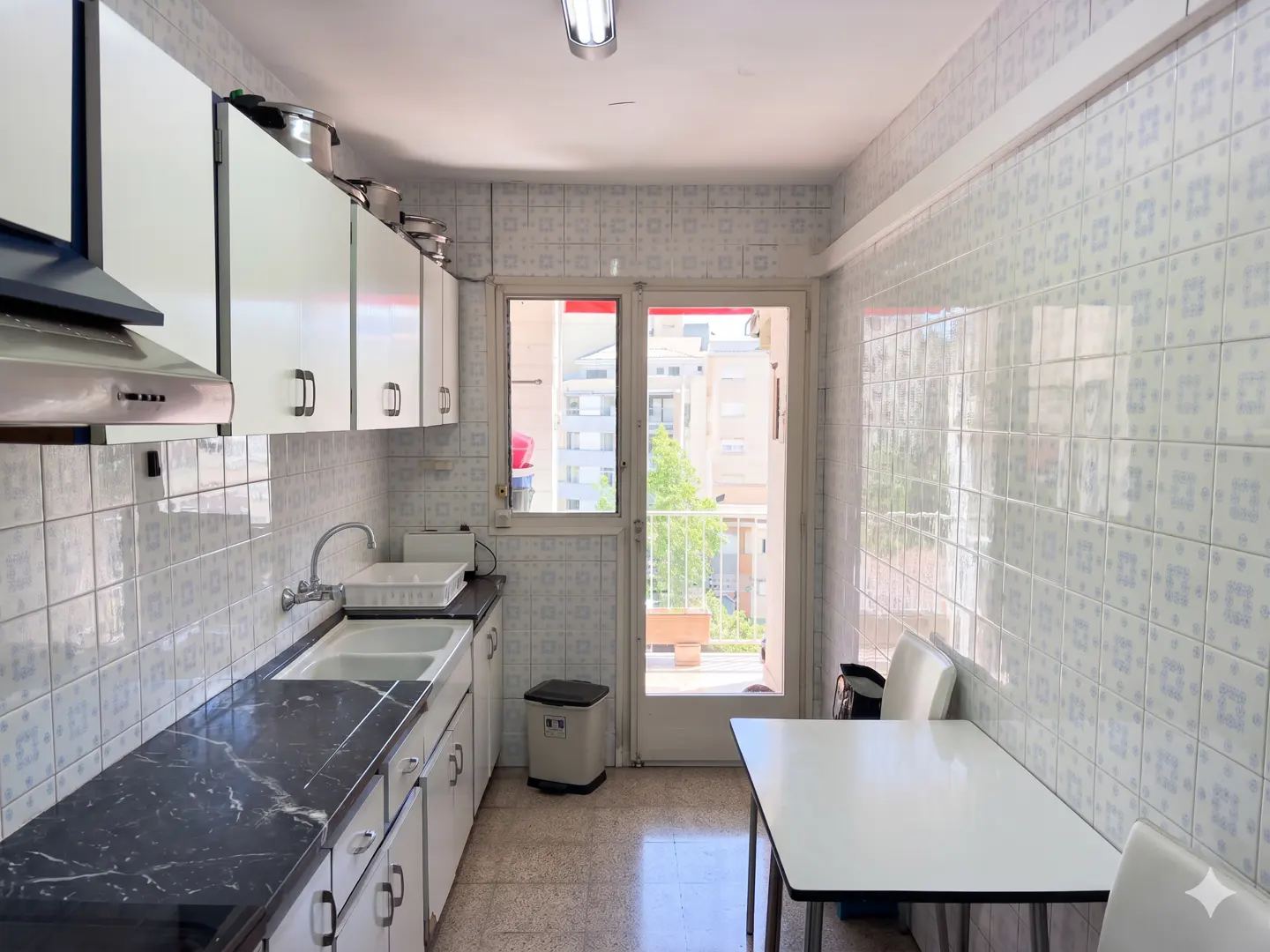 A narrow kitchen with white cabinets, black marble countertops, and a white table near a window.