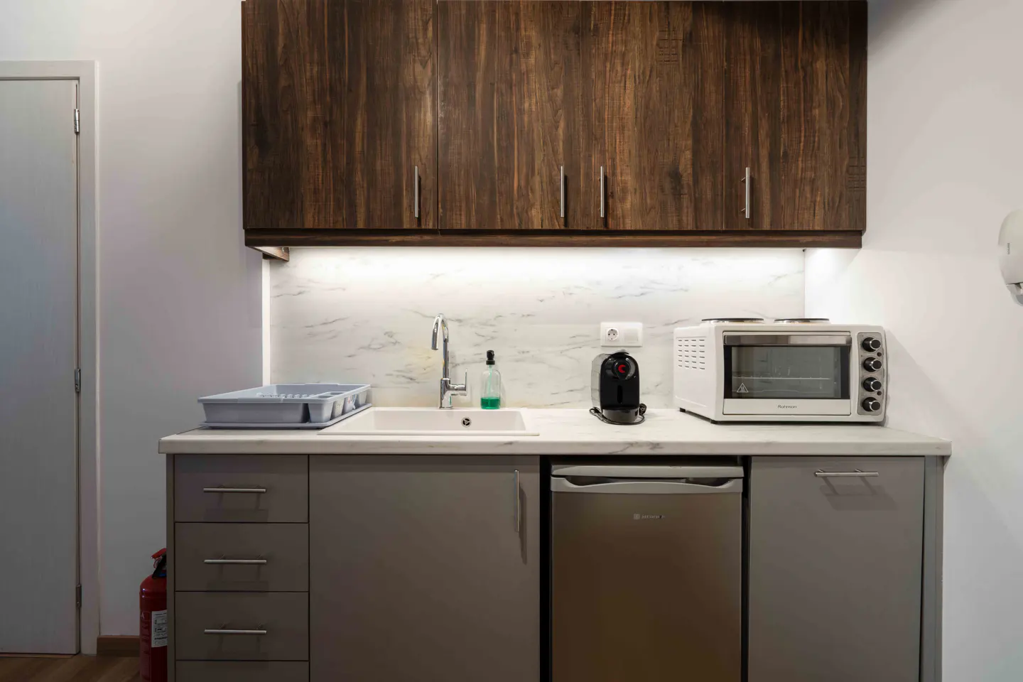 A compact kitchen area with gray cabinets, a white countertop, and dark wood upper cabinets. Appliances include a mini-fridge, microwave, and coffee maker.