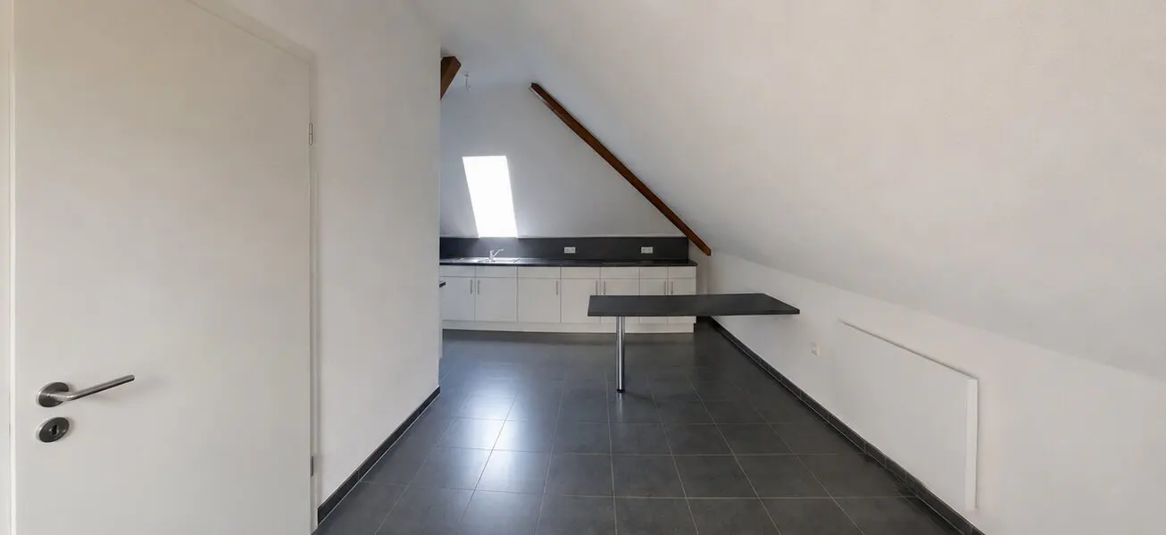 Attic apartment with white walls, gray tile floor, and a small kitchen area with white cabinets. A black table is attached to the wall.