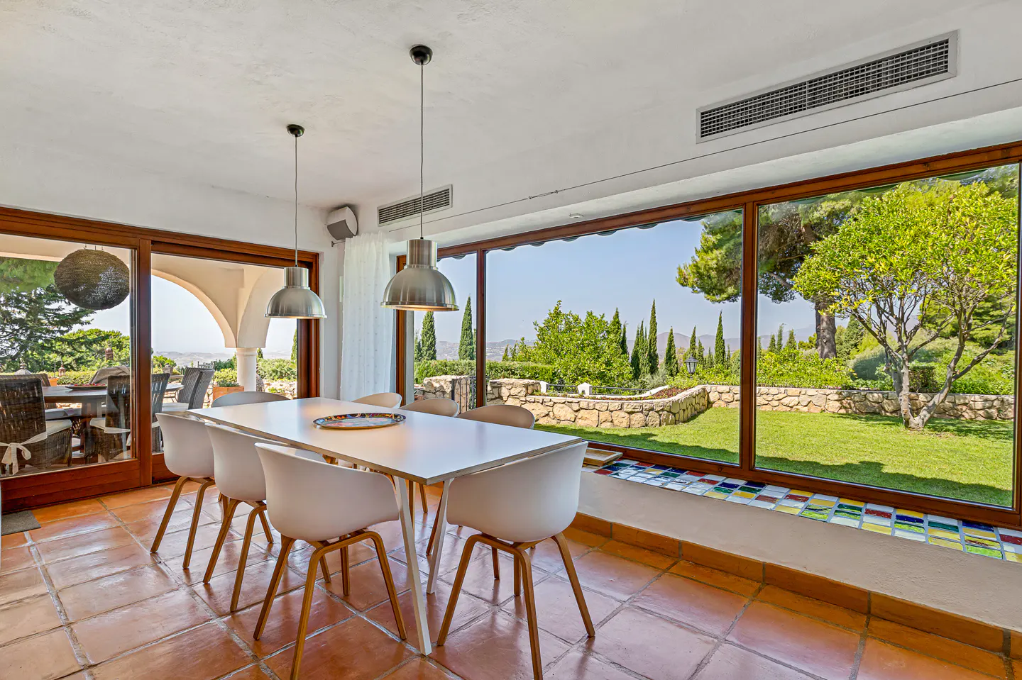 Bright dining room with white table, chairs, and pendant lights. Large windows overlook a green lawn and garden. Brown tile floor.