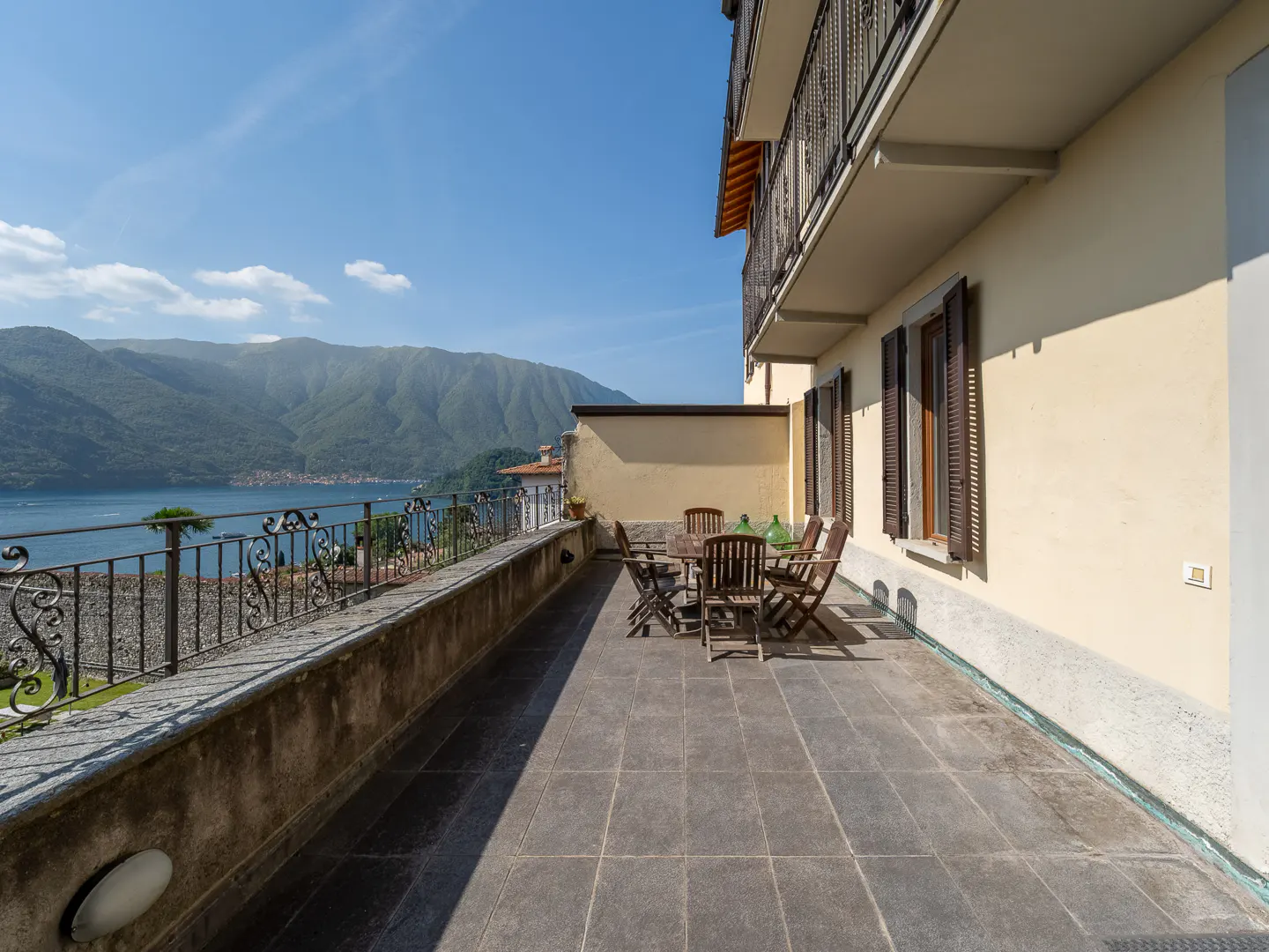 Spacious terrace with wooden dining set overlooking a scenic lake and mountains under a clear blue sky.