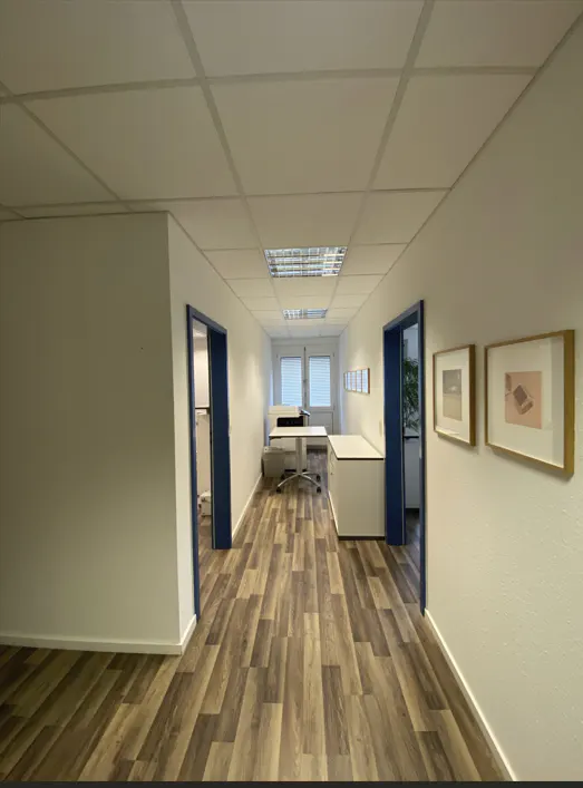 A bright office hallway with wood-look flooring, white walls, blue door frames, and two framed pictures on the right wall.