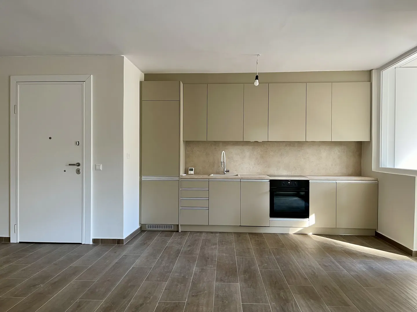 A modern kitchen with light brown cabinets, a black oven, and a stainless steel sink. A white door is on the left, and wood-look tile covers the floor.