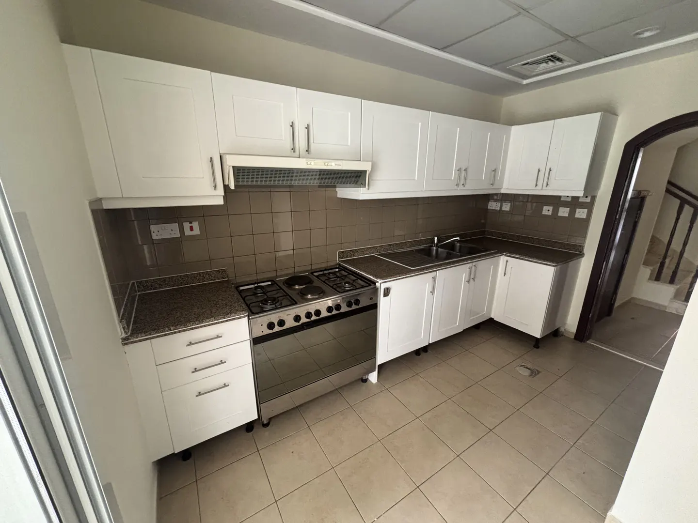 Bright kitchen with white cabinets, stainless steel stove, and tiled floor. A staircase is visible through an arched doorway.