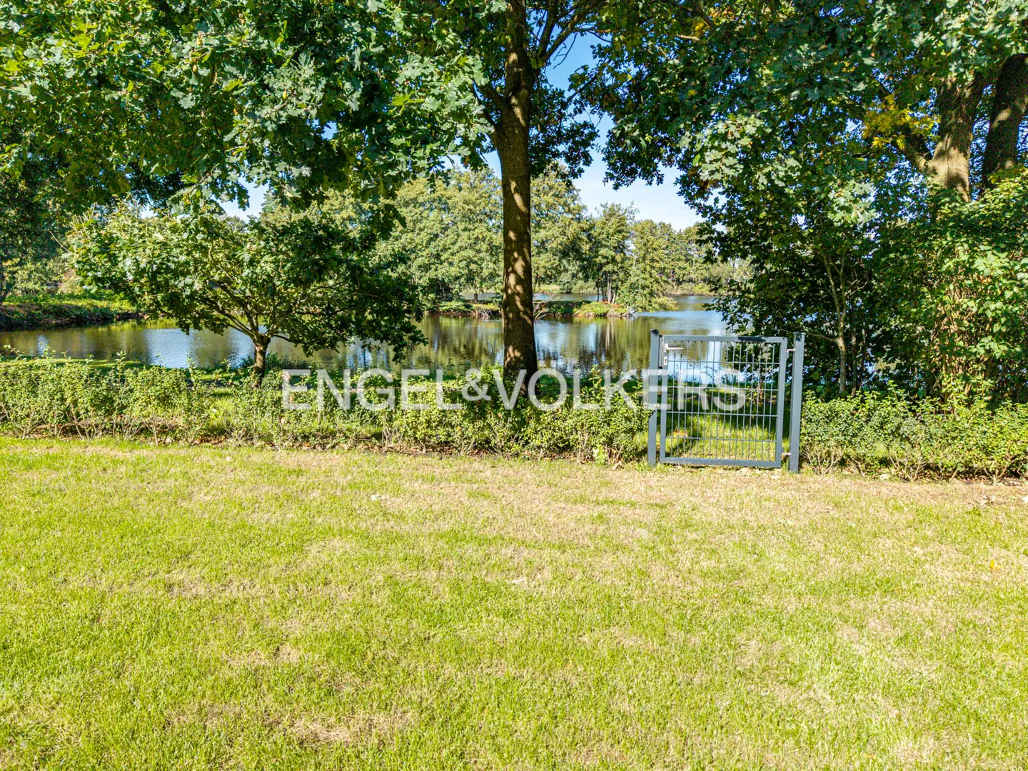 A green lawn leads to a small metal gate opening toward a calm pond surrounded by lush trees on a sunny day.