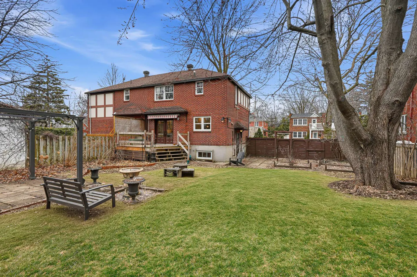 Backyard view of a two-story red brick house with a green lawn, patio furniture, and a large tree on a sunny day.