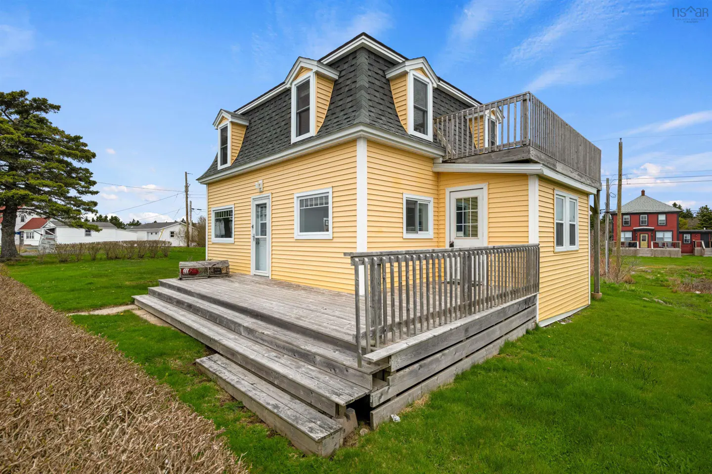 Yellow two-story house with a grey shingled roof, a large wooden deck, and a second-floor balcony set on a grassy lawn.