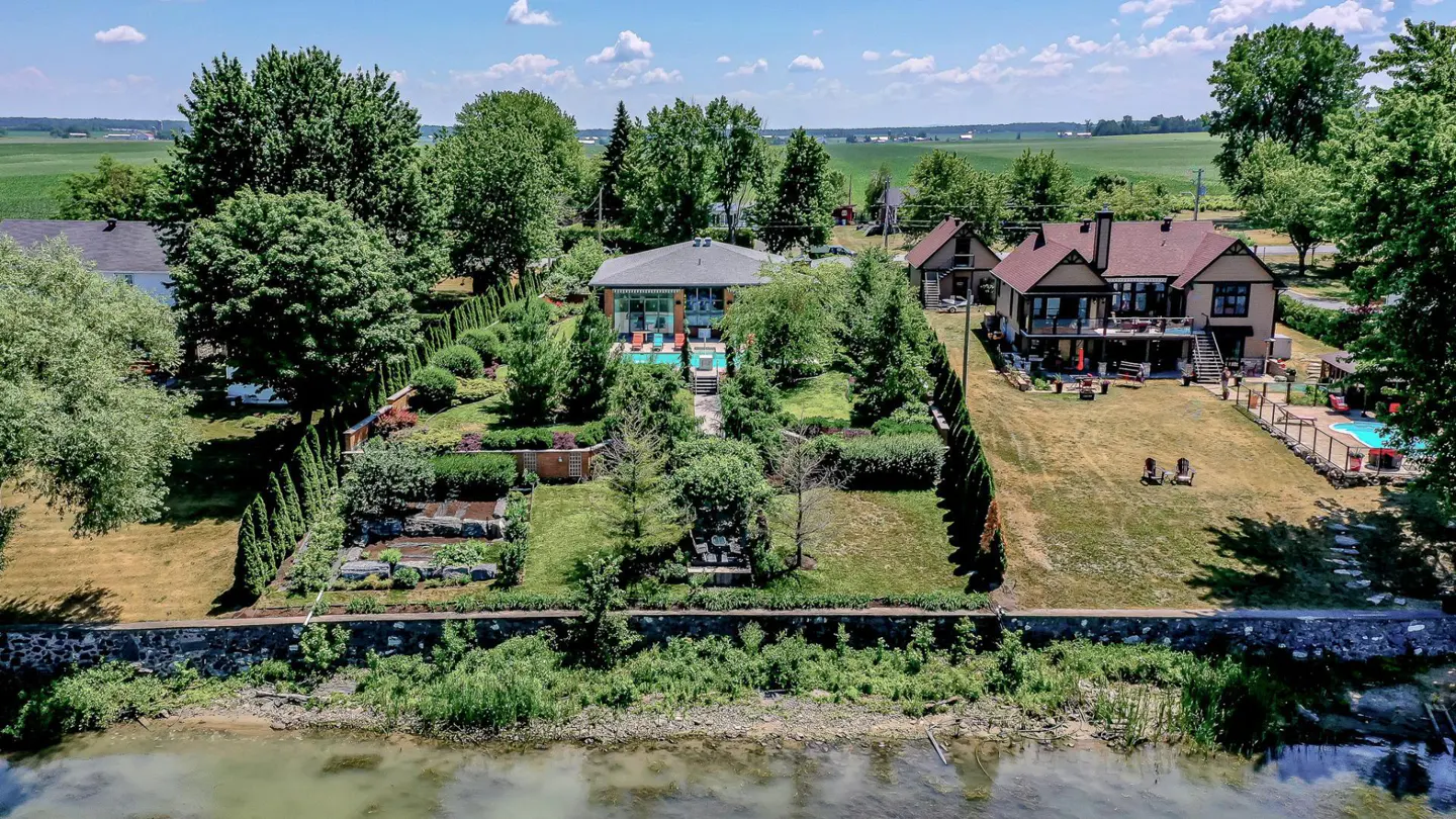 Aerial view of two waterfront homes with pools, manicured lawns, and lush greenery on a sunny day.